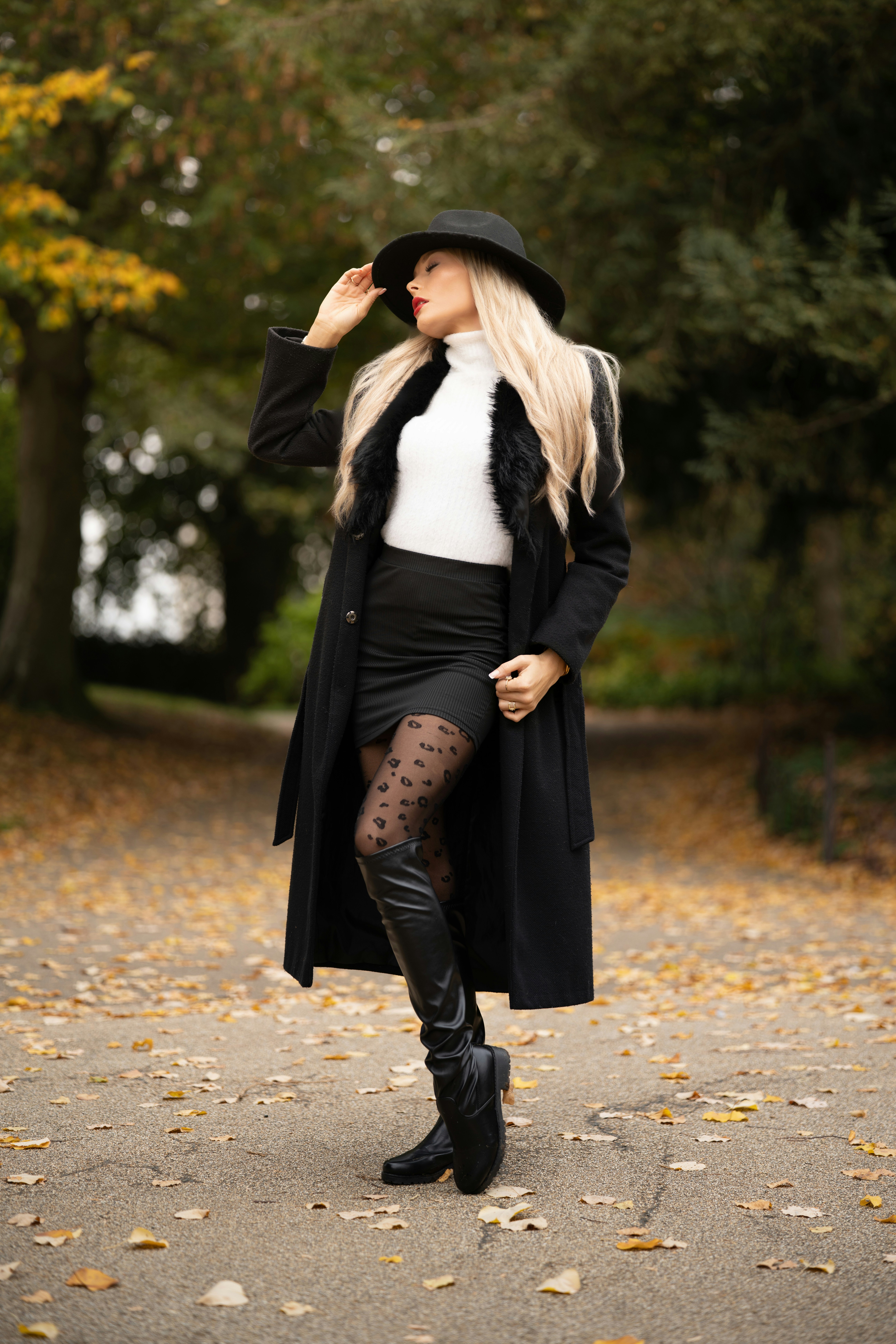 Woman in a black coat and fedora stands among autumn leaves.
