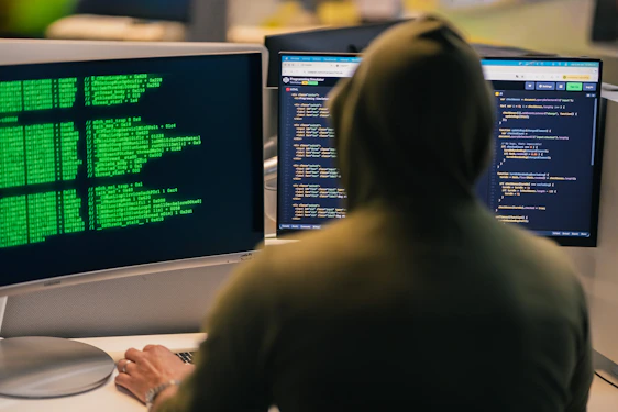 Hacker in hoodie working on multiple computer screens