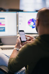 Man using smartphone in front of computer monitors.