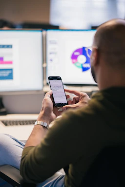 Man using smartphone in front of computer monitors.
