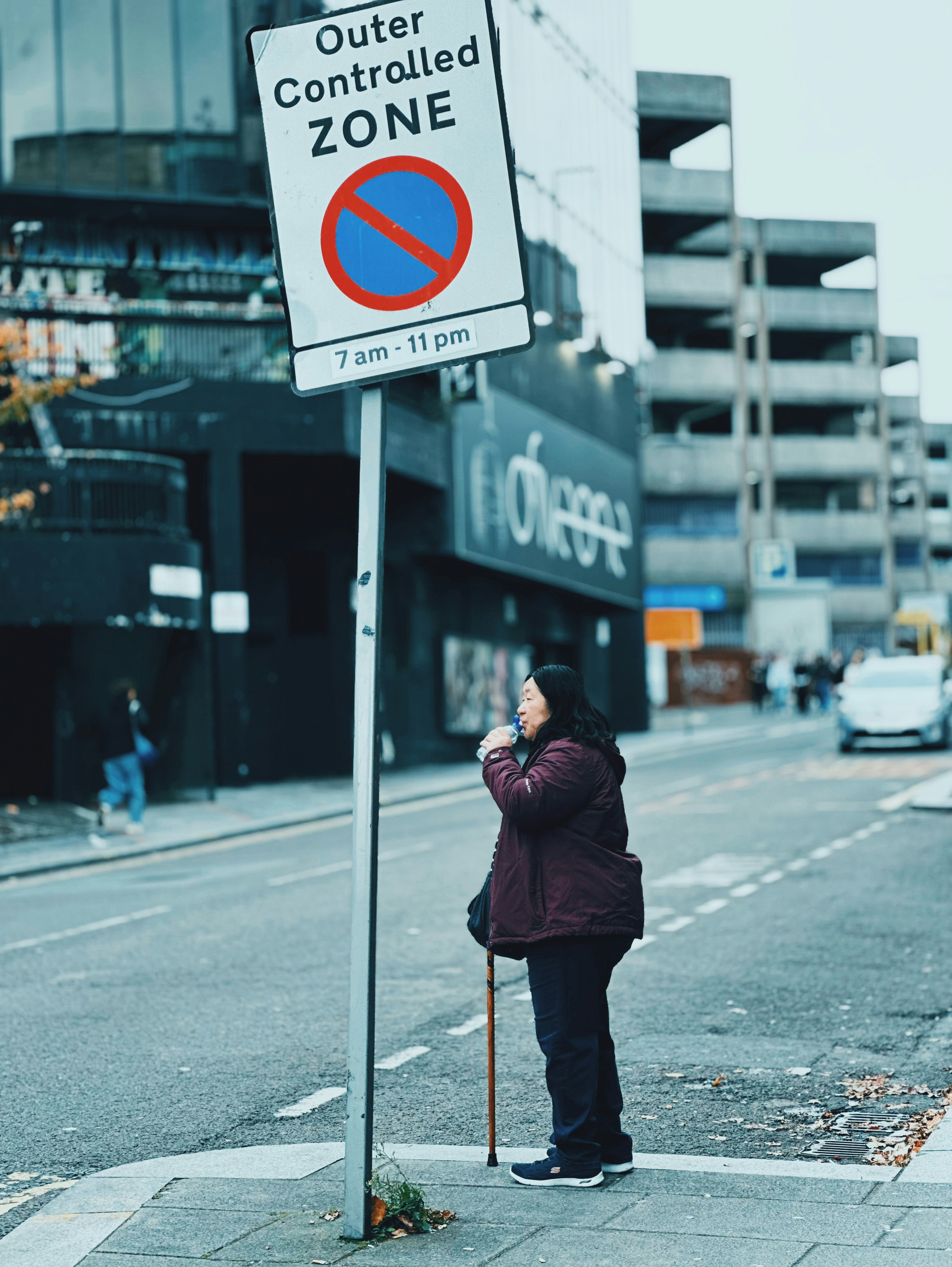 Une femme avec une canne attend au coin de la rue près du panneau.