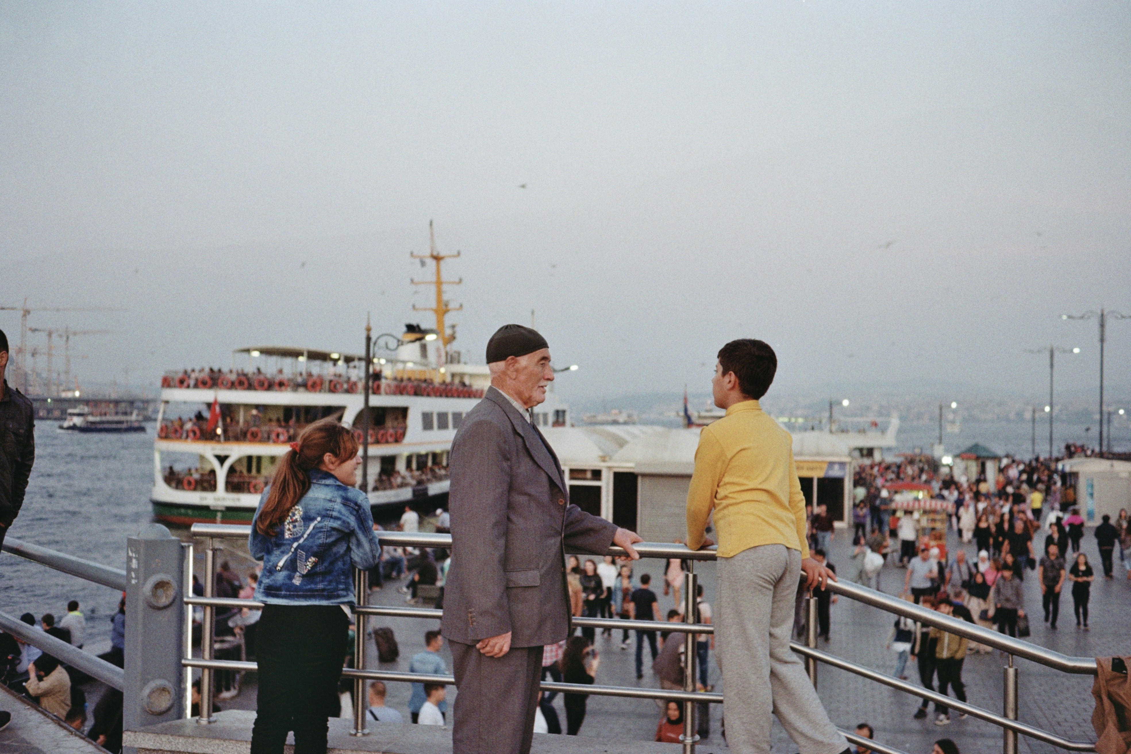 People gathered by a ferry terminal with boats on water.