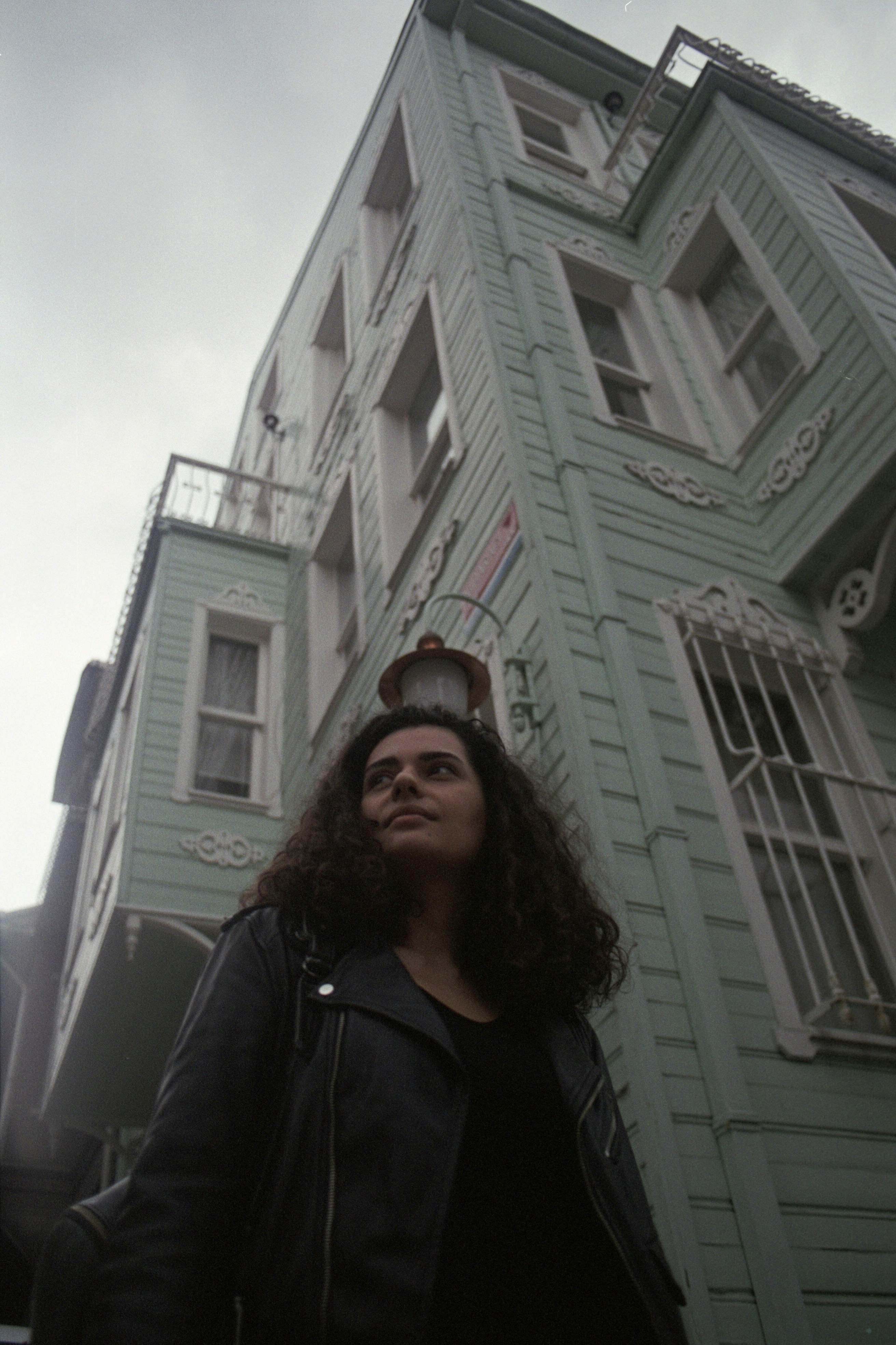 Woman in leather jacket stands before pale green building