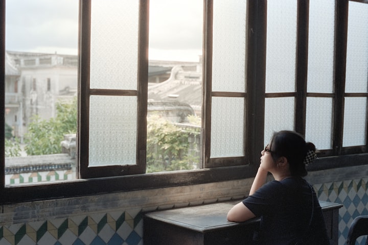 Profile of a person at a window, looking out toward distant buildings in soft daylight