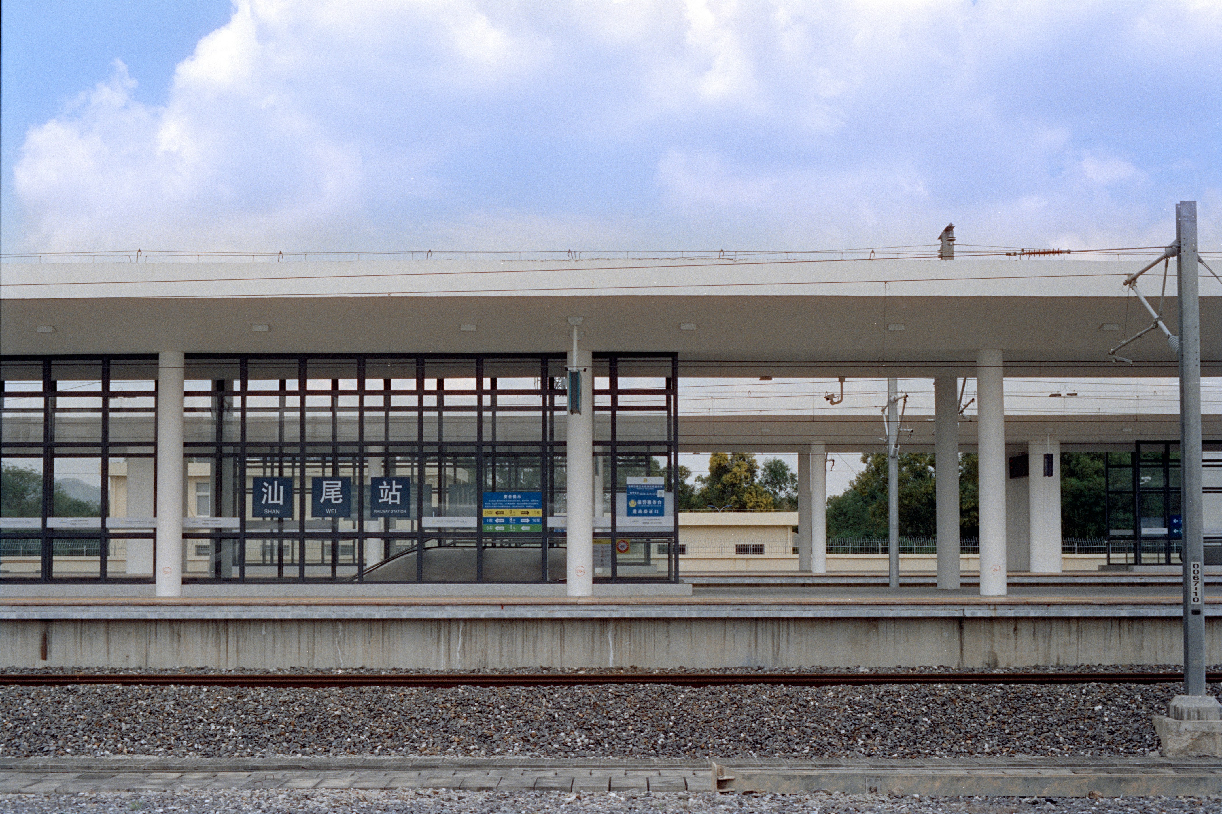 A detailed architectural rendering of a modern railway station exterior, showcasing sleek design, green spaces, and integrated commercial areas.
