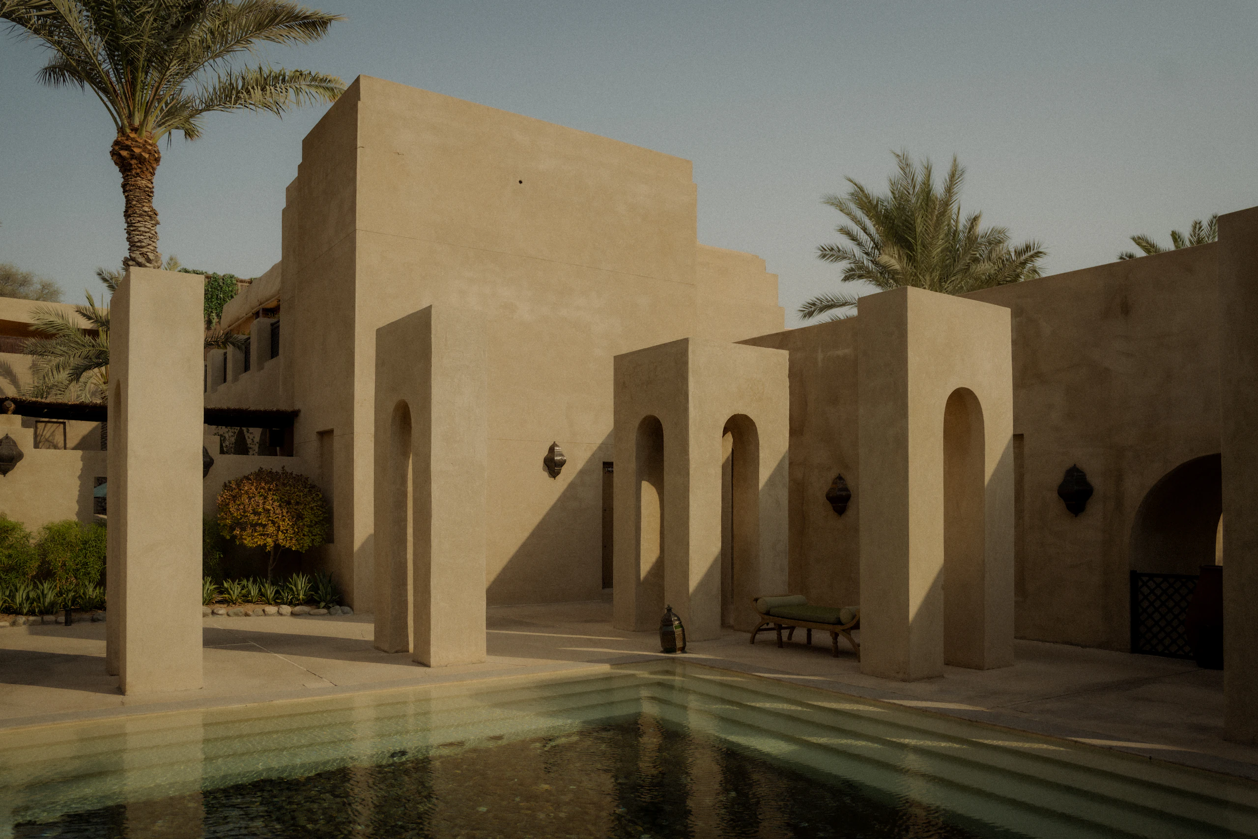 A sand-coloured arcade of slender arches reflected in a still pool at dusk, framed by date palms.