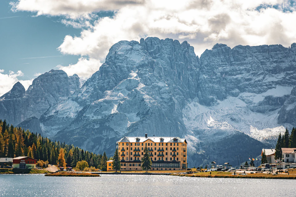 The image depicts a serene mountain landscape with a large, snow-capped peak dominating the scene. The mountain is surrounded by lush green trees and shrubs, creating a beautiful contrast between nature's elements. In the foreground, there is a tranquil lake reflecting the surrounding scenery. On the right side of the image, several buildings can be seen, suggesting that this location may have human habitation or serve as a popular tourist destination.

In the center of the image, there is a large yellow building with multiple stories and balconies, standing out against the natural backdrop. The sky above is clear blue with fluffy white clouds, adding to the overall beauty of the scene.