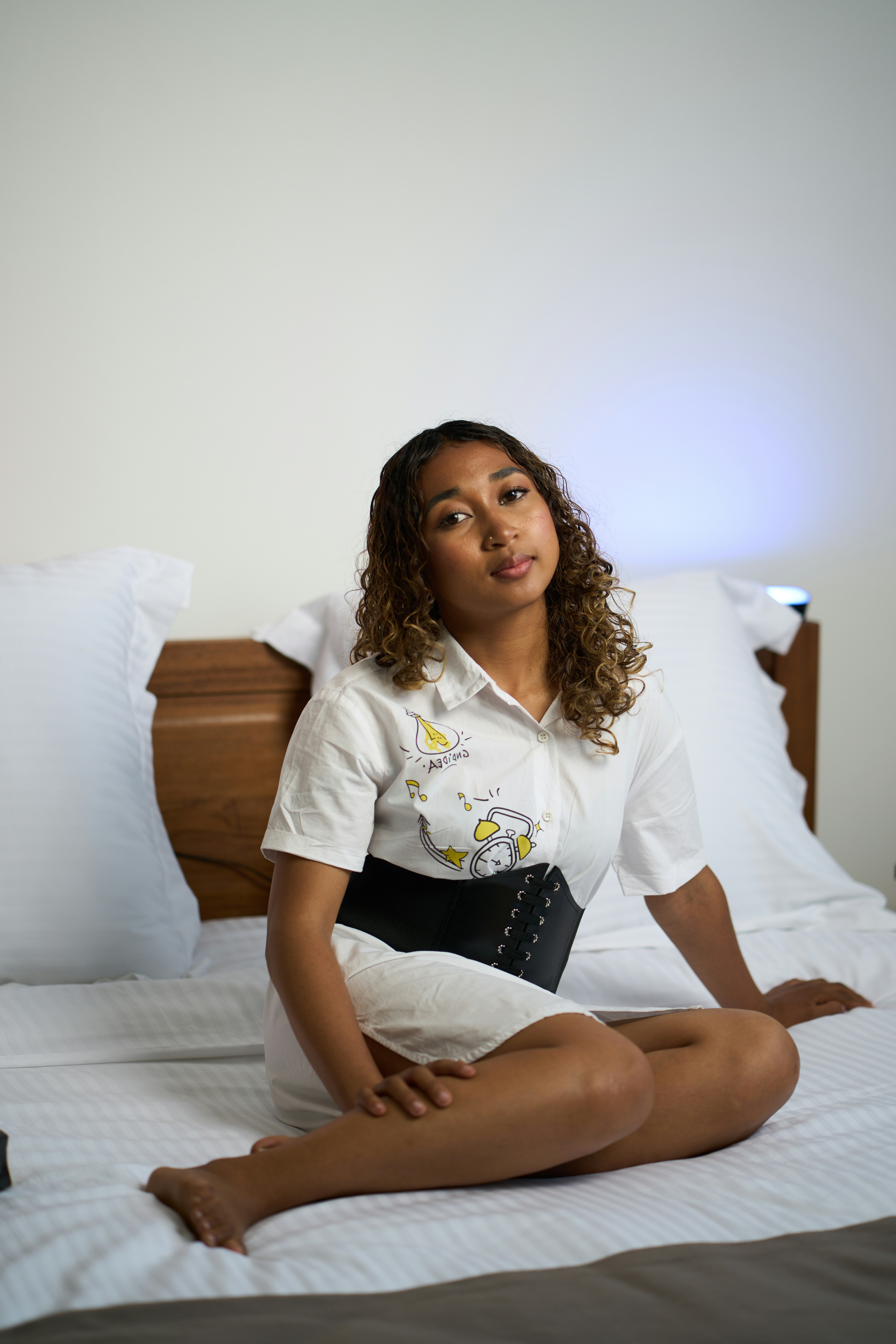 Young woman sits on a bed wearing a white dress.