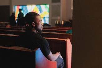 Man sitting in church pews looking towards stained glass.
