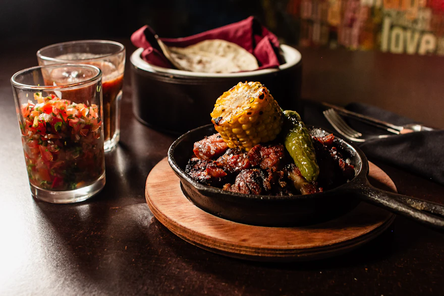 A cast iron skillet of grilled meat with roasted corn and a blistered chile, salsa in small glasses and a covered tortilla warmer