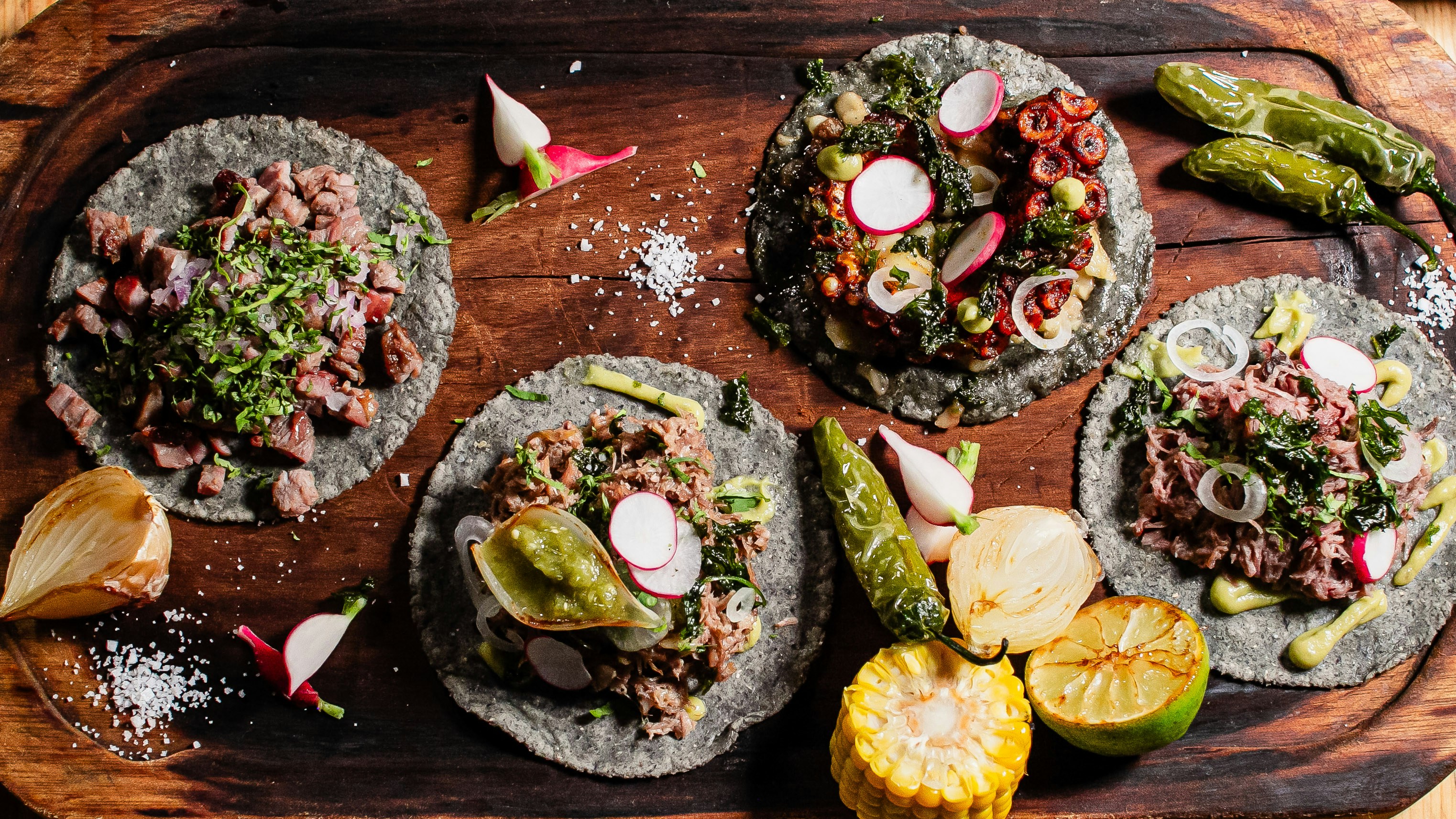 Four gourmet tacos with assorted toppings on wooden board