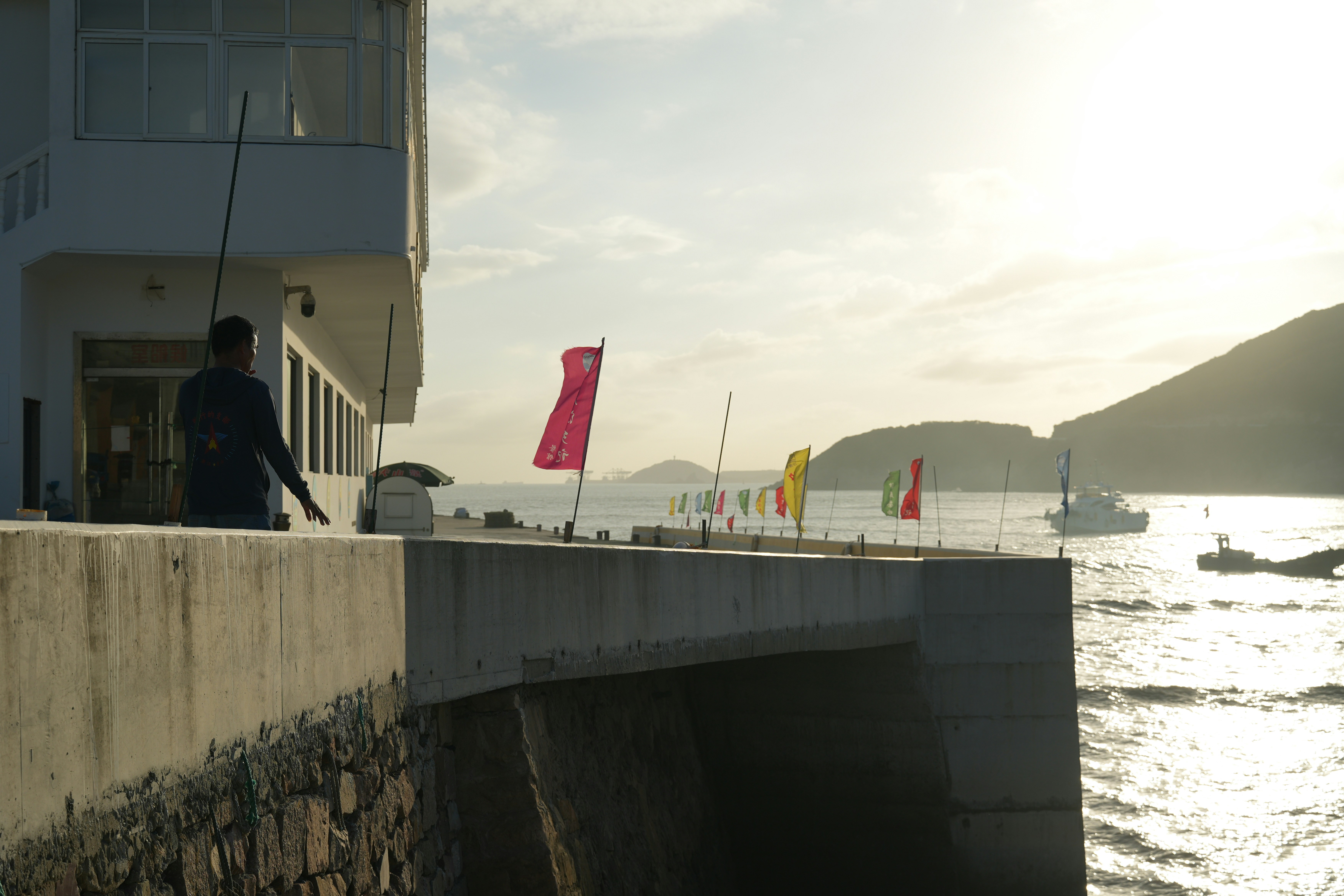 Man fishing from a pier with colorful flags and boats.