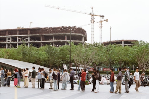 People stand in line near construction site with cranes.