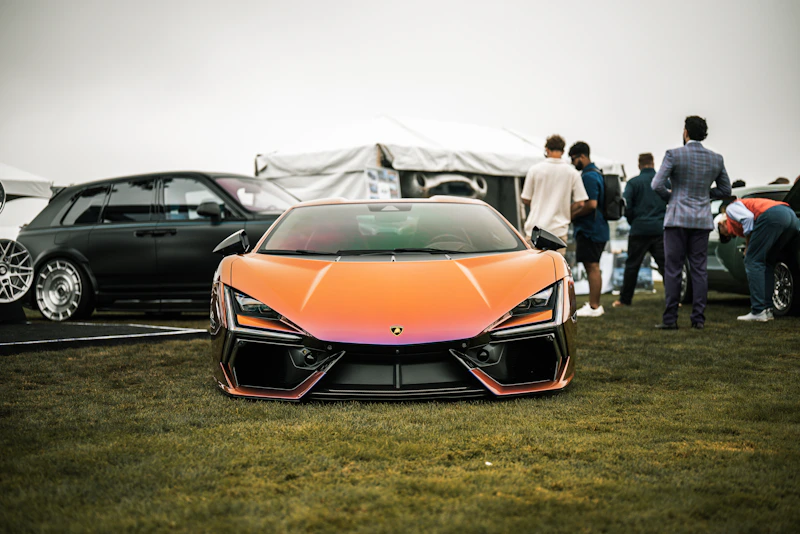 An orange sports car parked on grass with people.