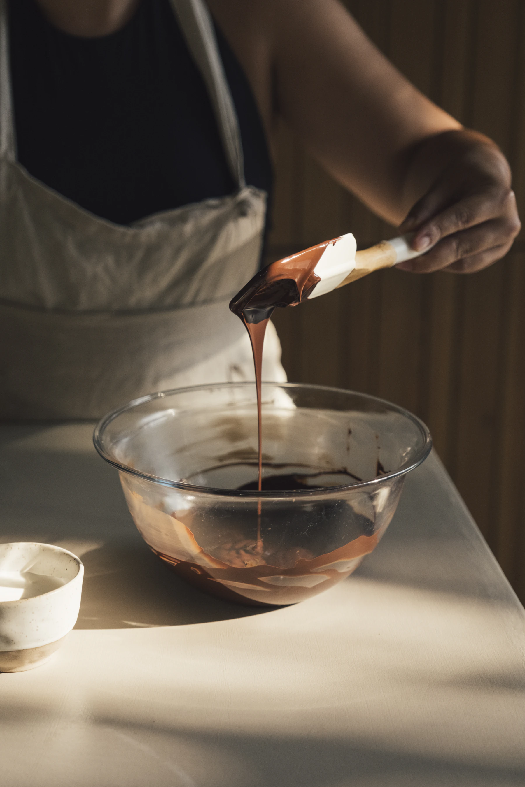 A cook in an apron lifts a white spatula of melted chocolate over a glass bowl