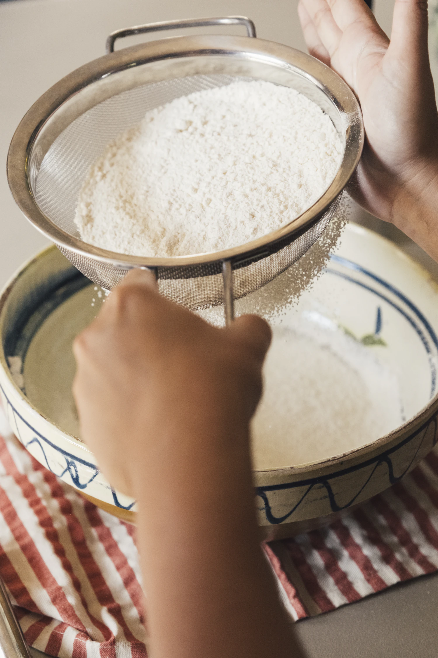 Two hands shaking flour through a round sieve into a bowl below