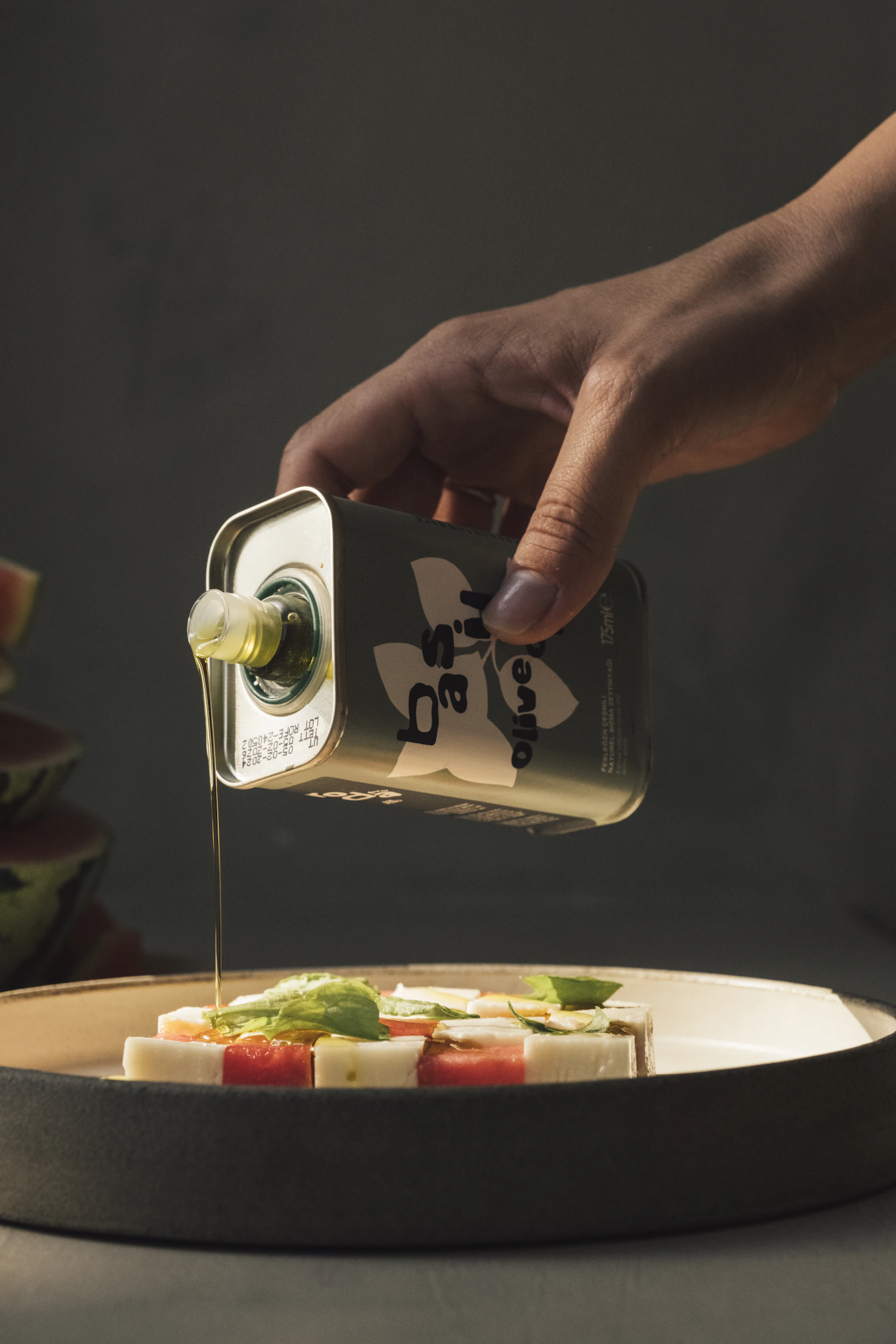 A hand pouring olive oil from a tin over tomato, mozzarella and basil