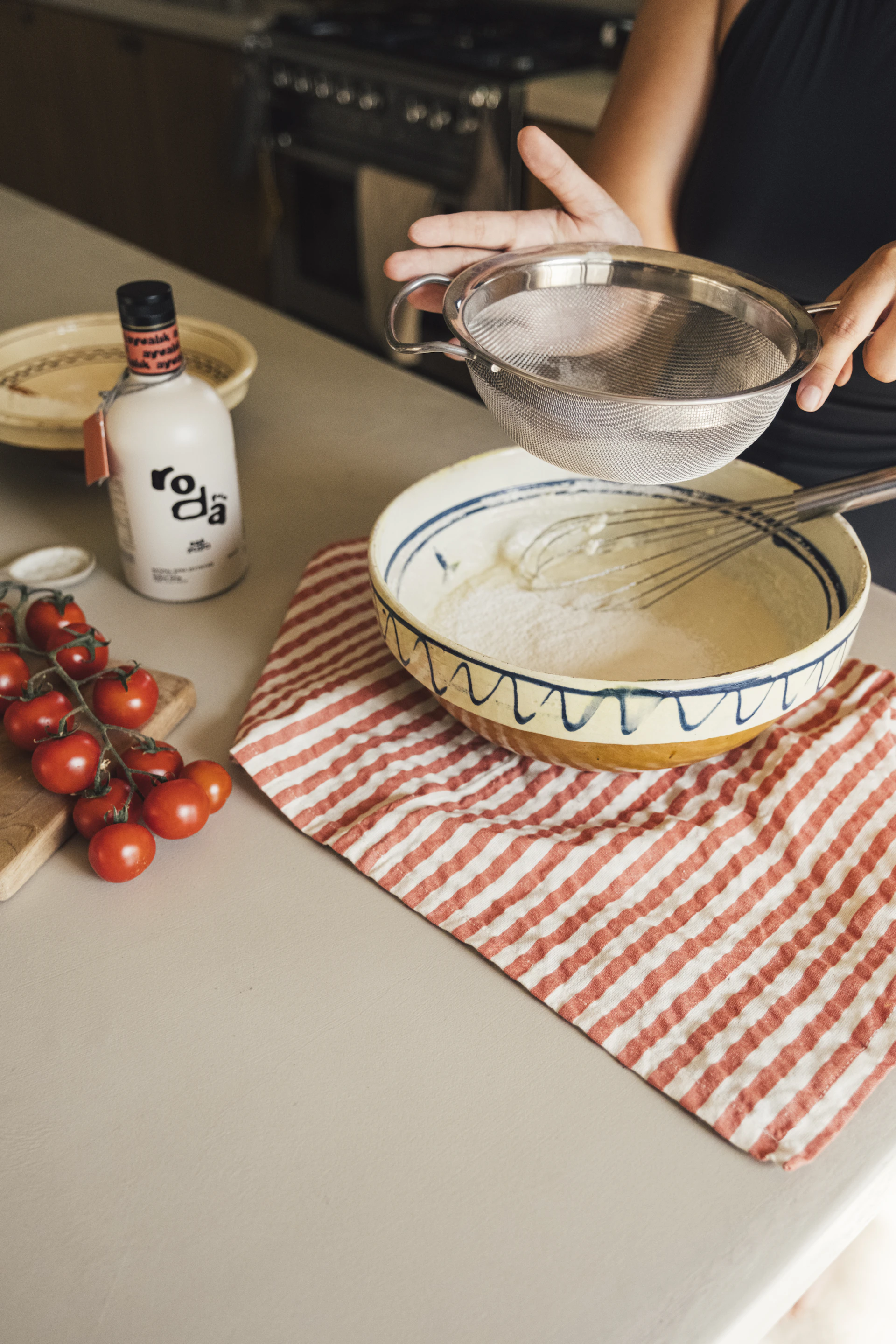 Person sifting flour into a bowl with tomatoes and bottle.
