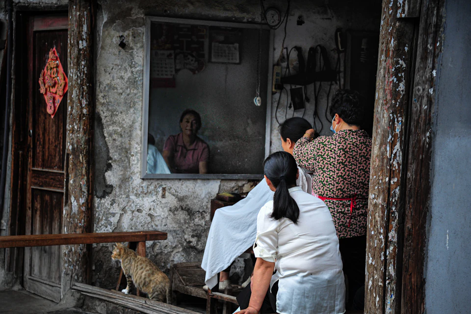 The image depicts a group of people gathered around a window, with one person holding a cat on their shoulder and another standing next to them. The scene takes place outside an old building, which appears to be made of stone or concrete. A wooden door can also be seen in the background.