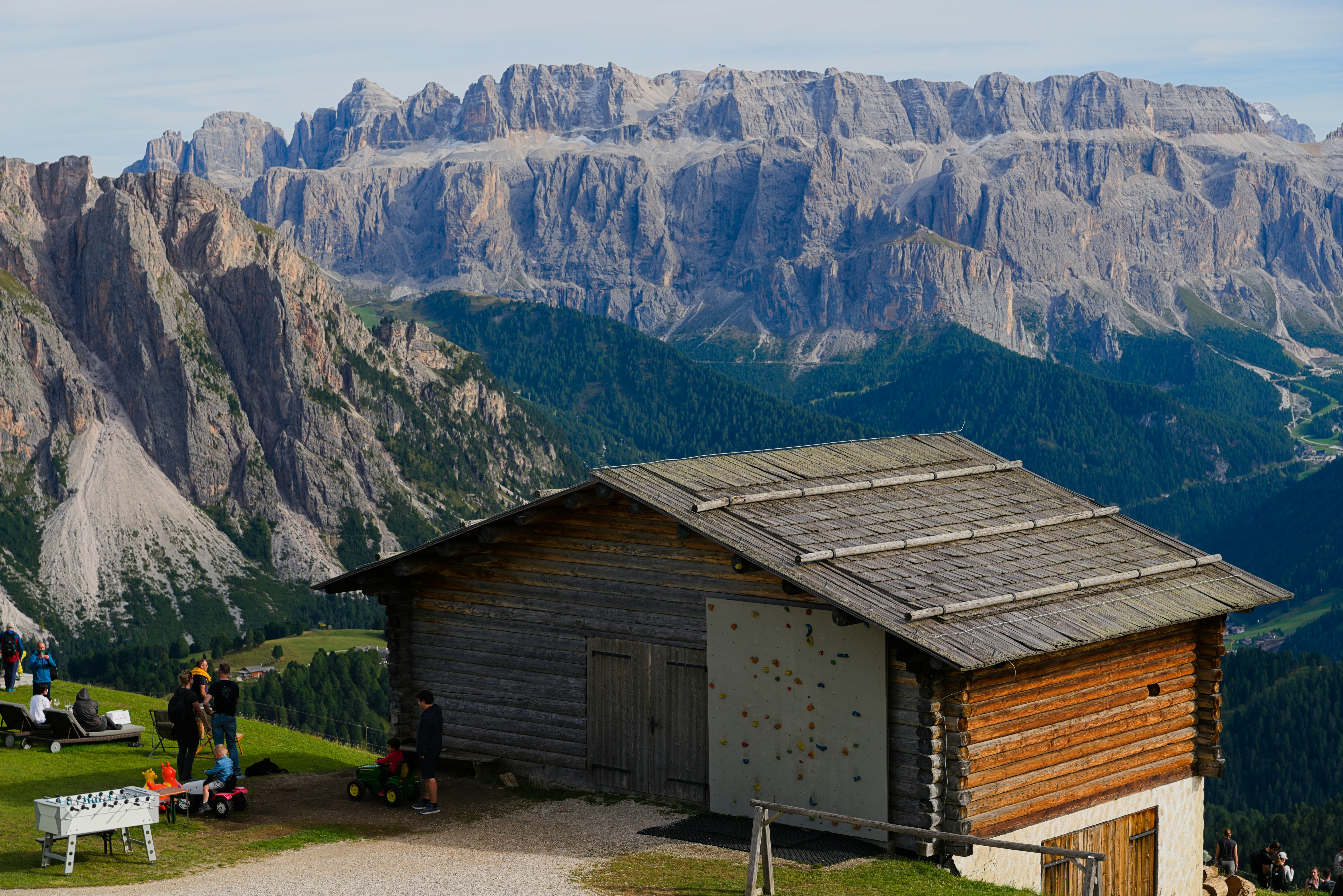 Wooden cabin with people on grassy hill and mountains.