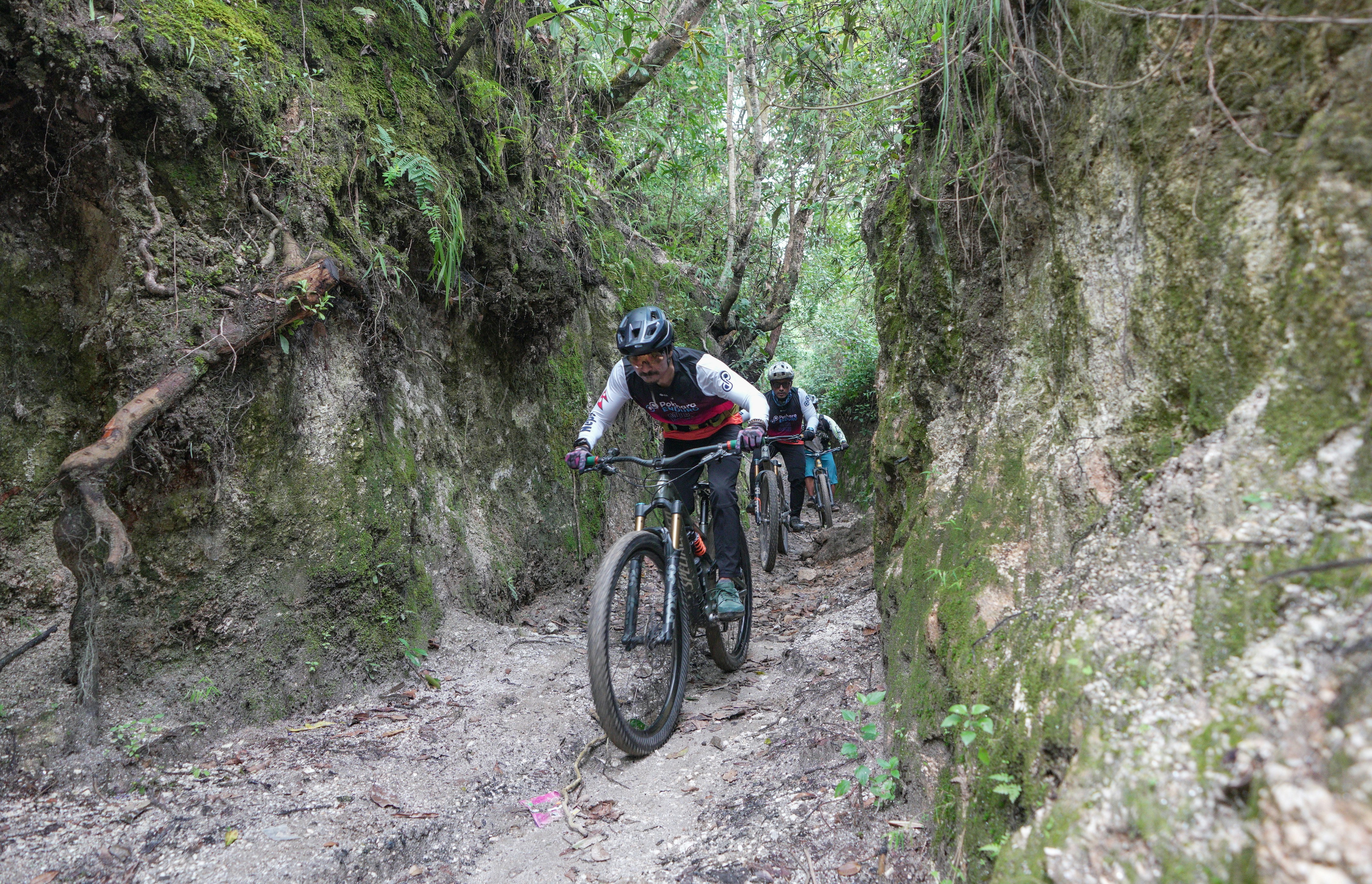 Cyclists ride through a narrow rocky forest path.