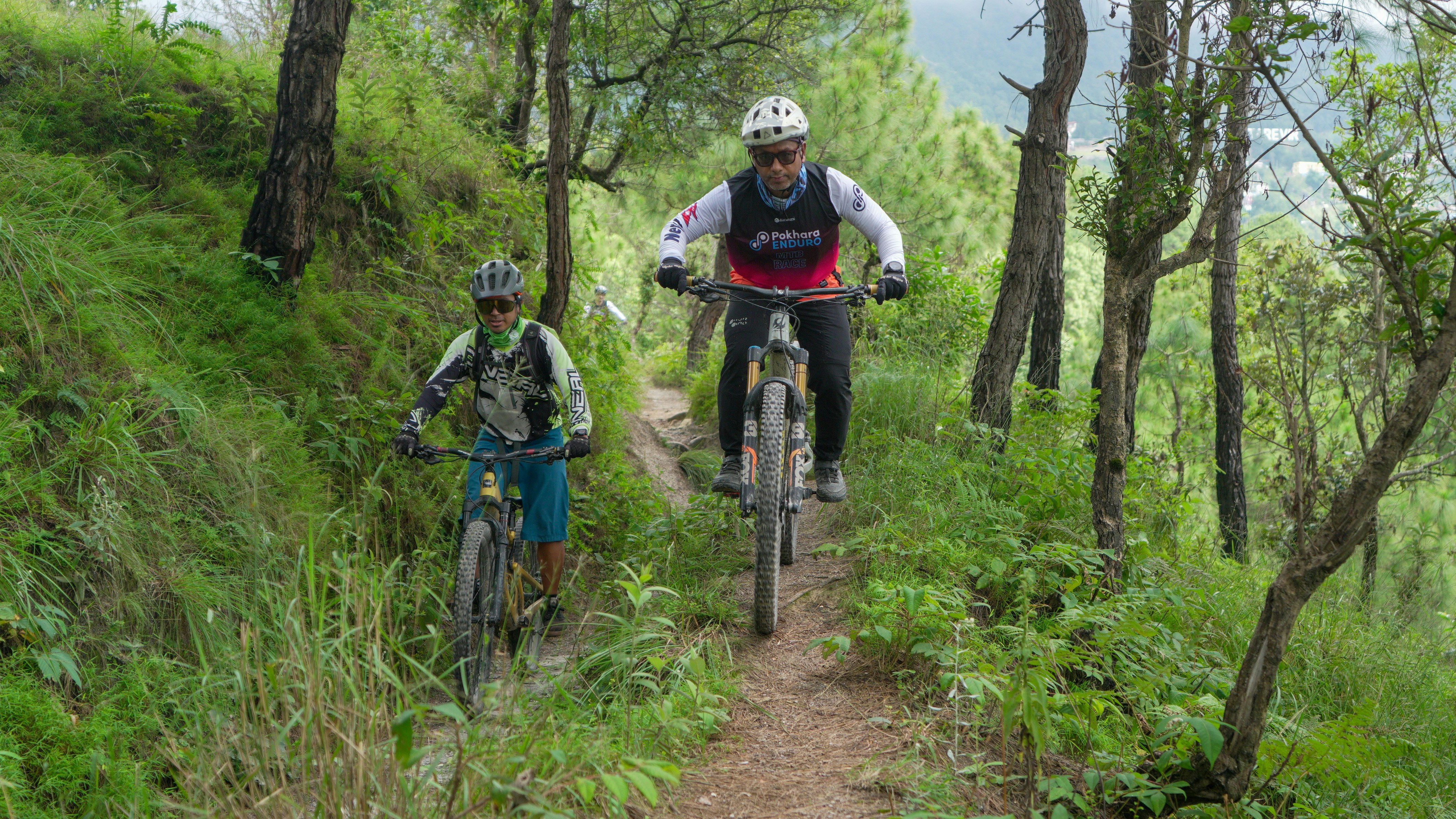 Two cyclists ride down a wooded mountain trail.