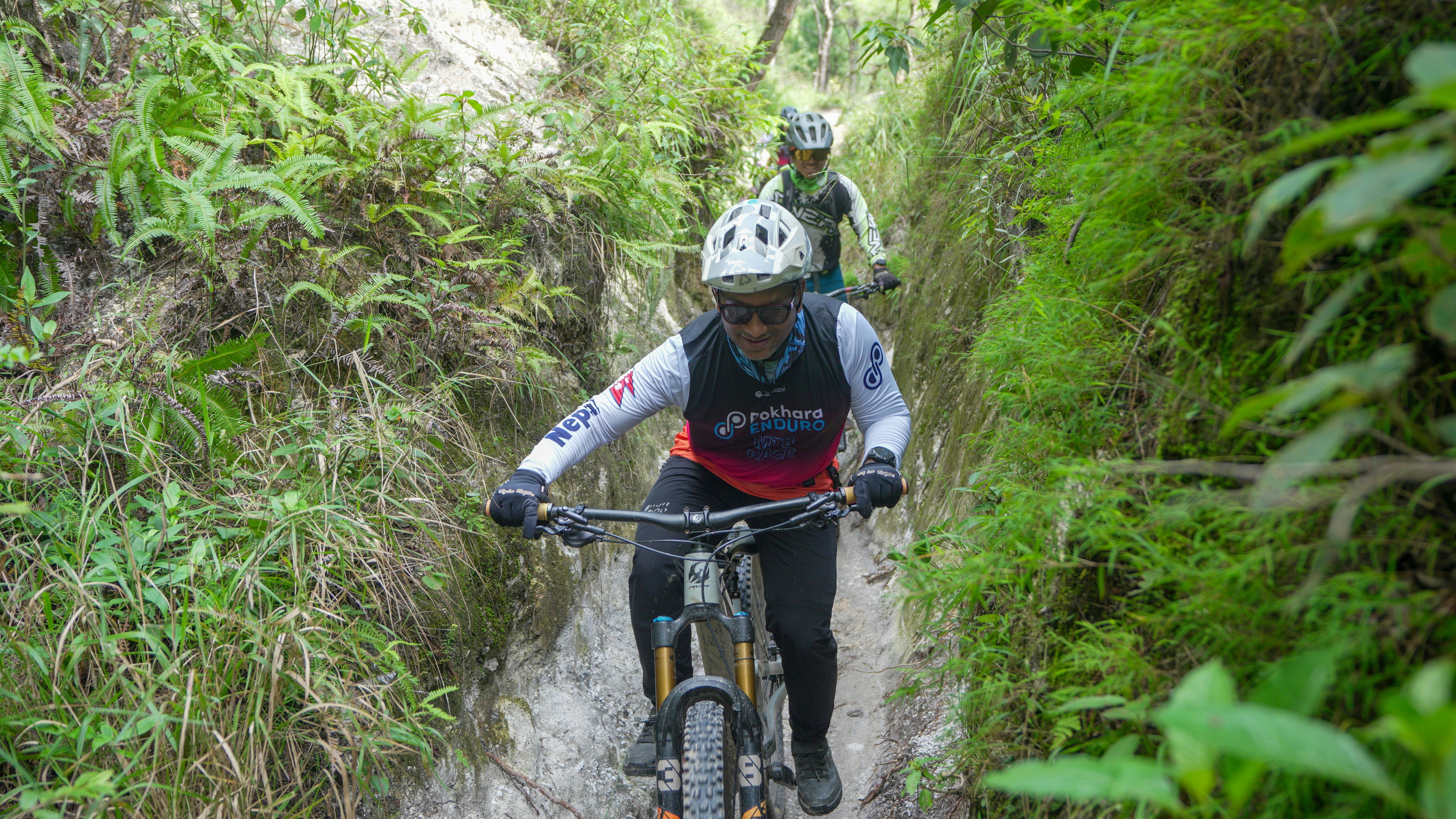 Two mountain bikers riding down a narrow, overgrown trail.