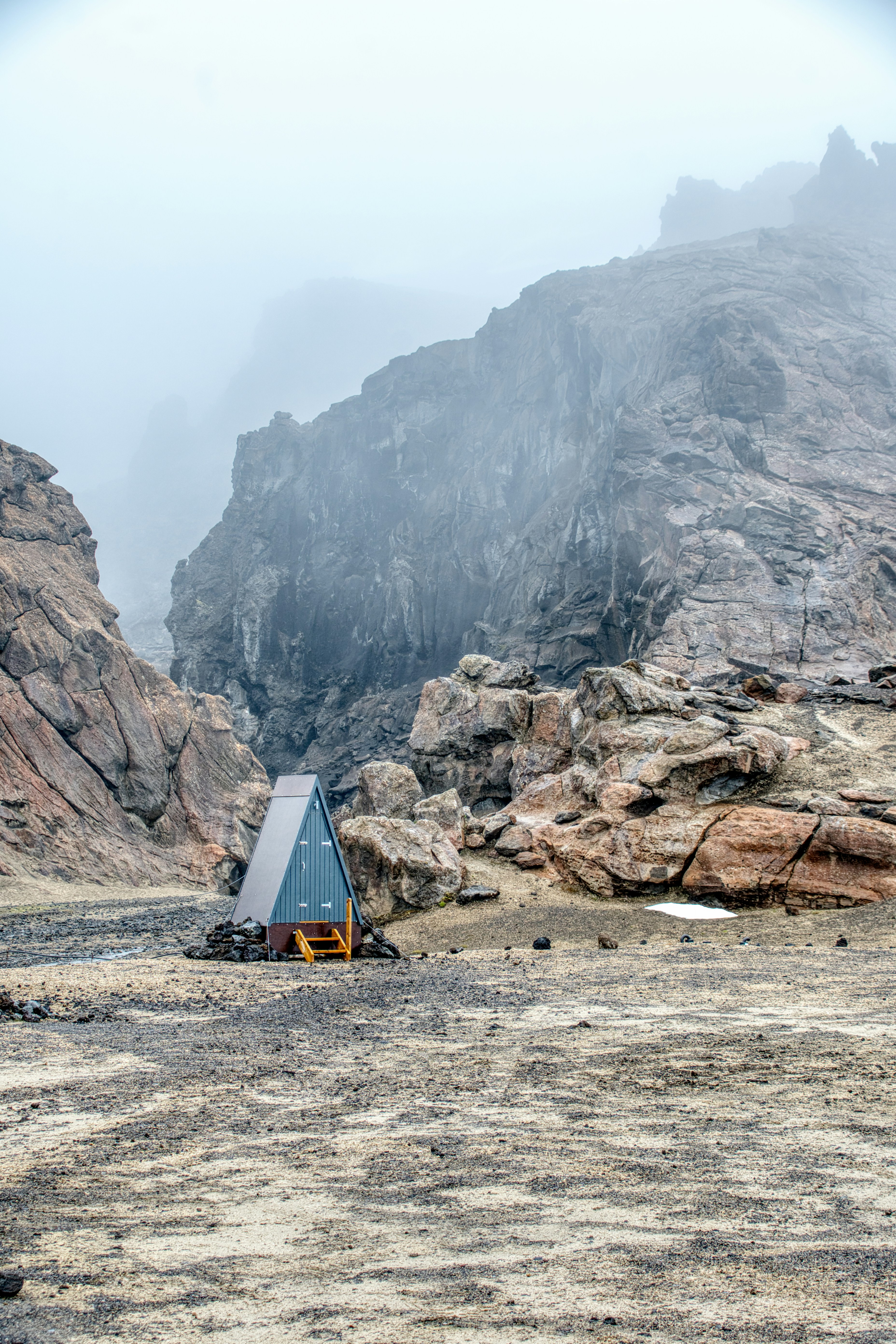 Piccolo rifugio con struttura ad A in un paesaggio montano roccioso e nebbioso.