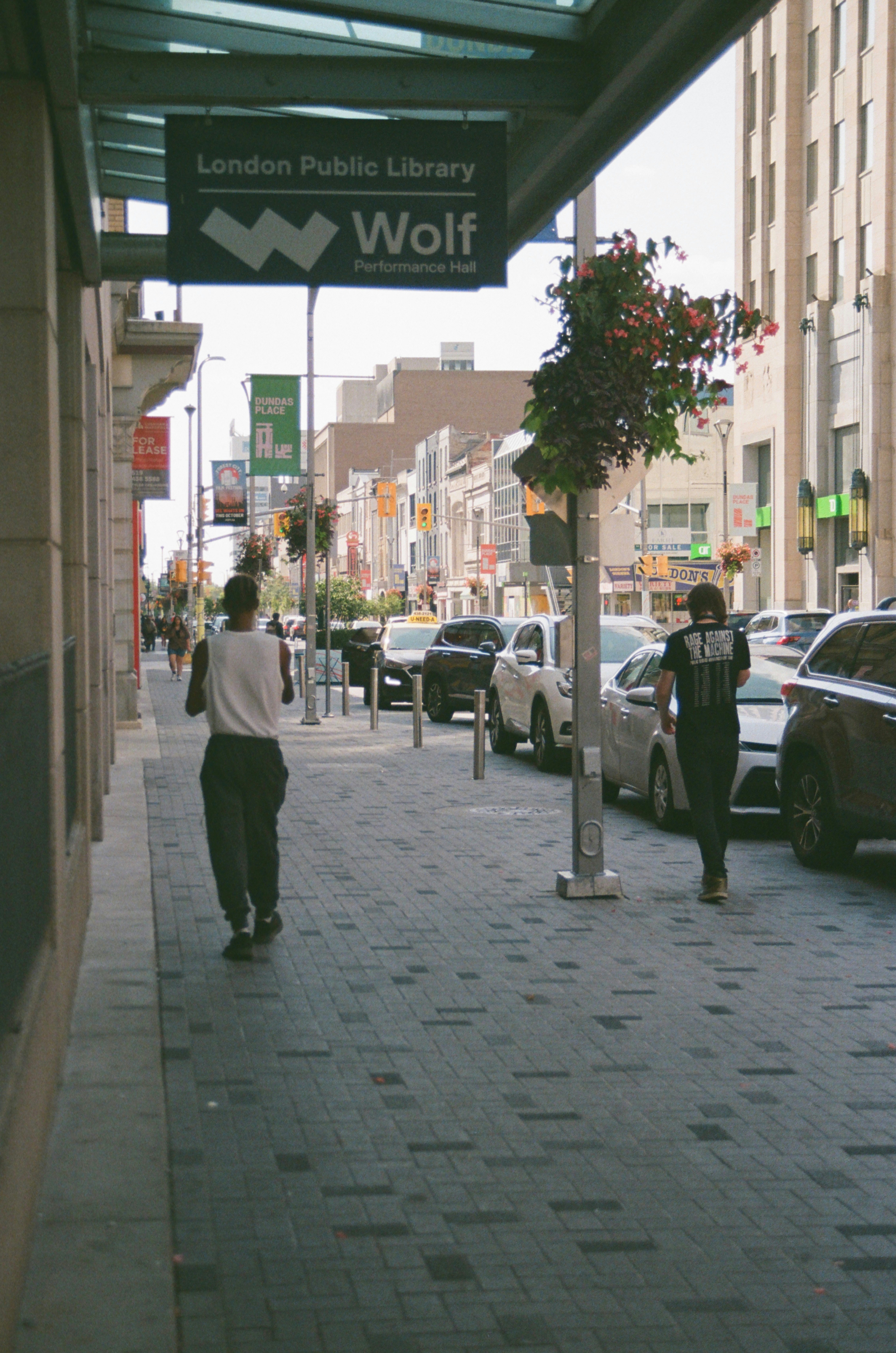 People walk down a city sidewalk beside parked cars.