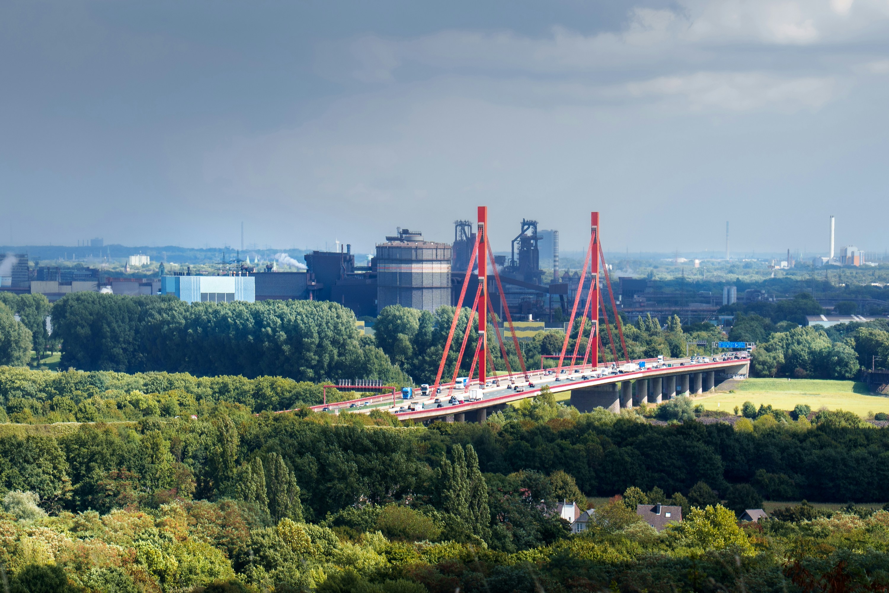 Modern red bridge spans across green landscape with industrial background