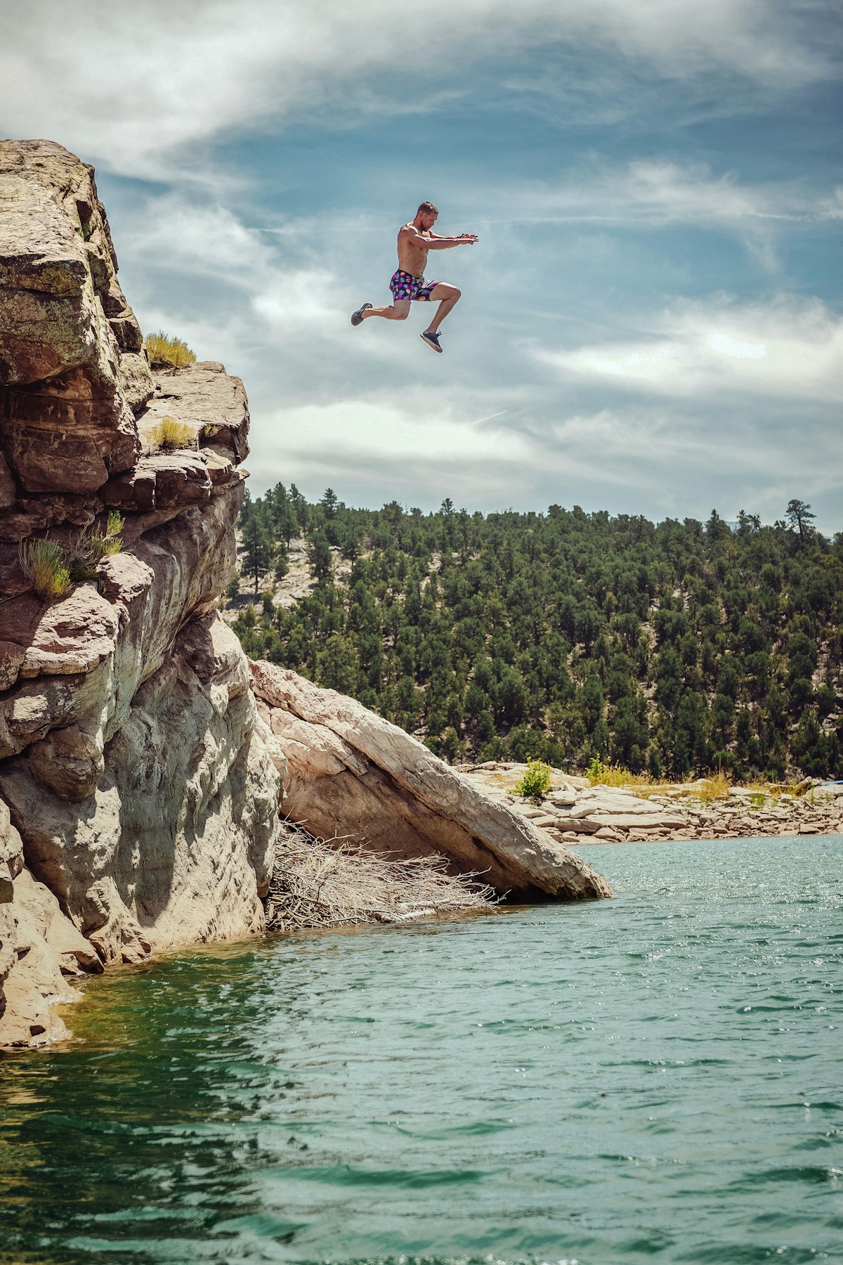 A man leaping off a rocky cliff into a turquoise lake, surrounded by pine forest