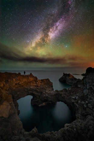 Man stands on rocky shore under starry night sky.