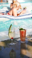 Two cocktails sit by a pool with people relaxing.