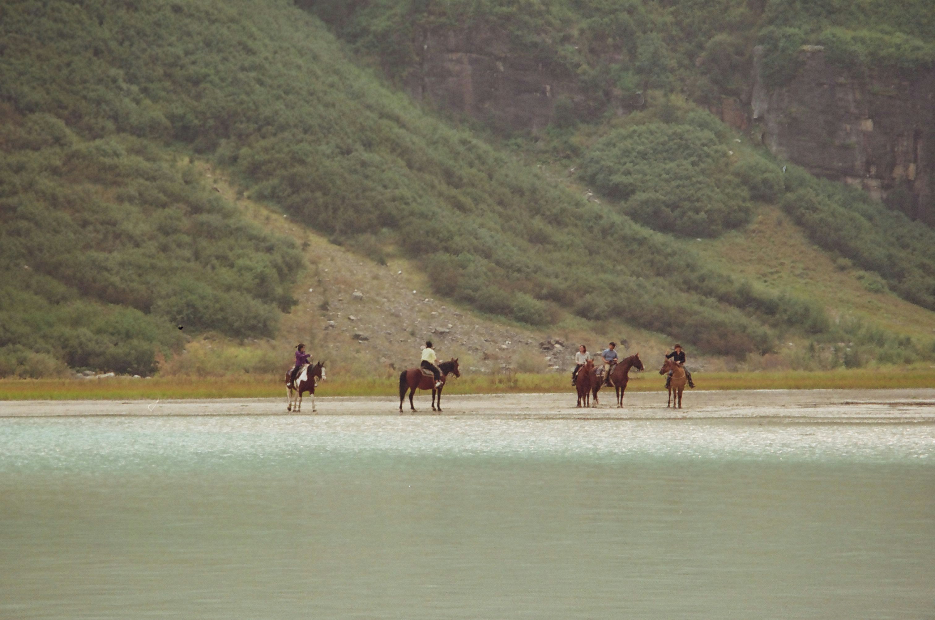 People on horseback riding along a calm body of water.
