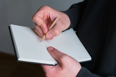 Person writing in a blank notebook with pencil.
