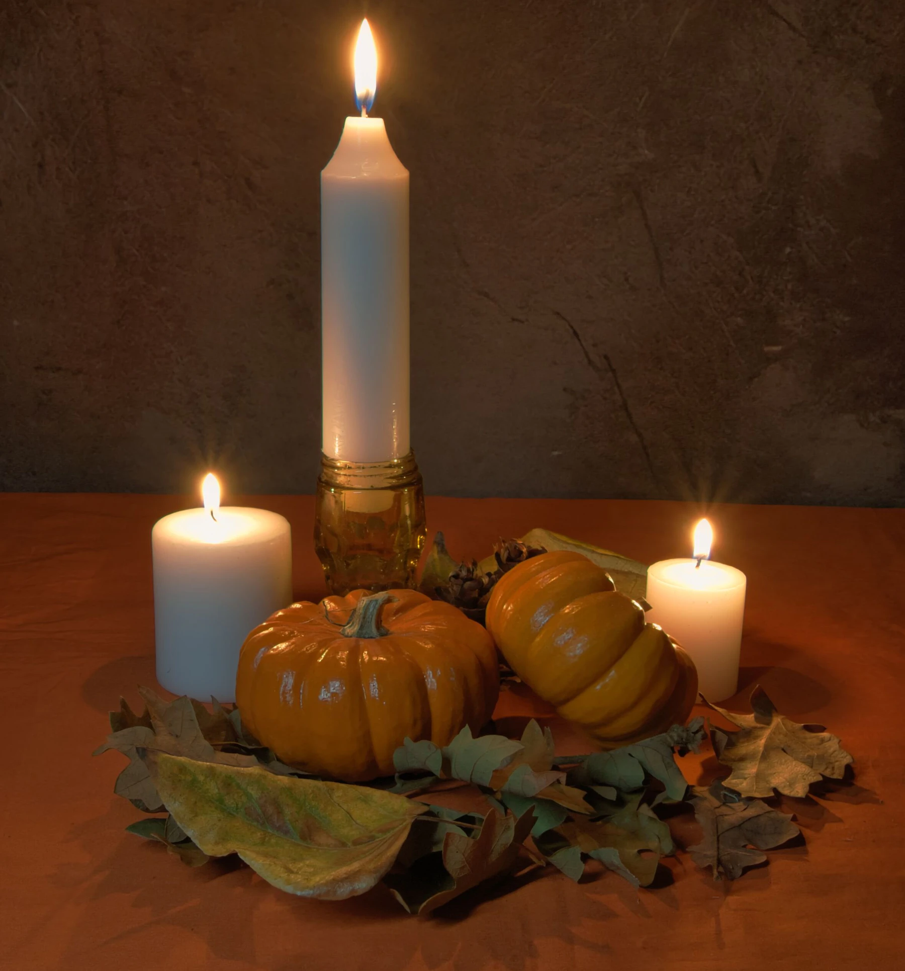 Three lit candles and small pumpkins surrounded by autumn leaves