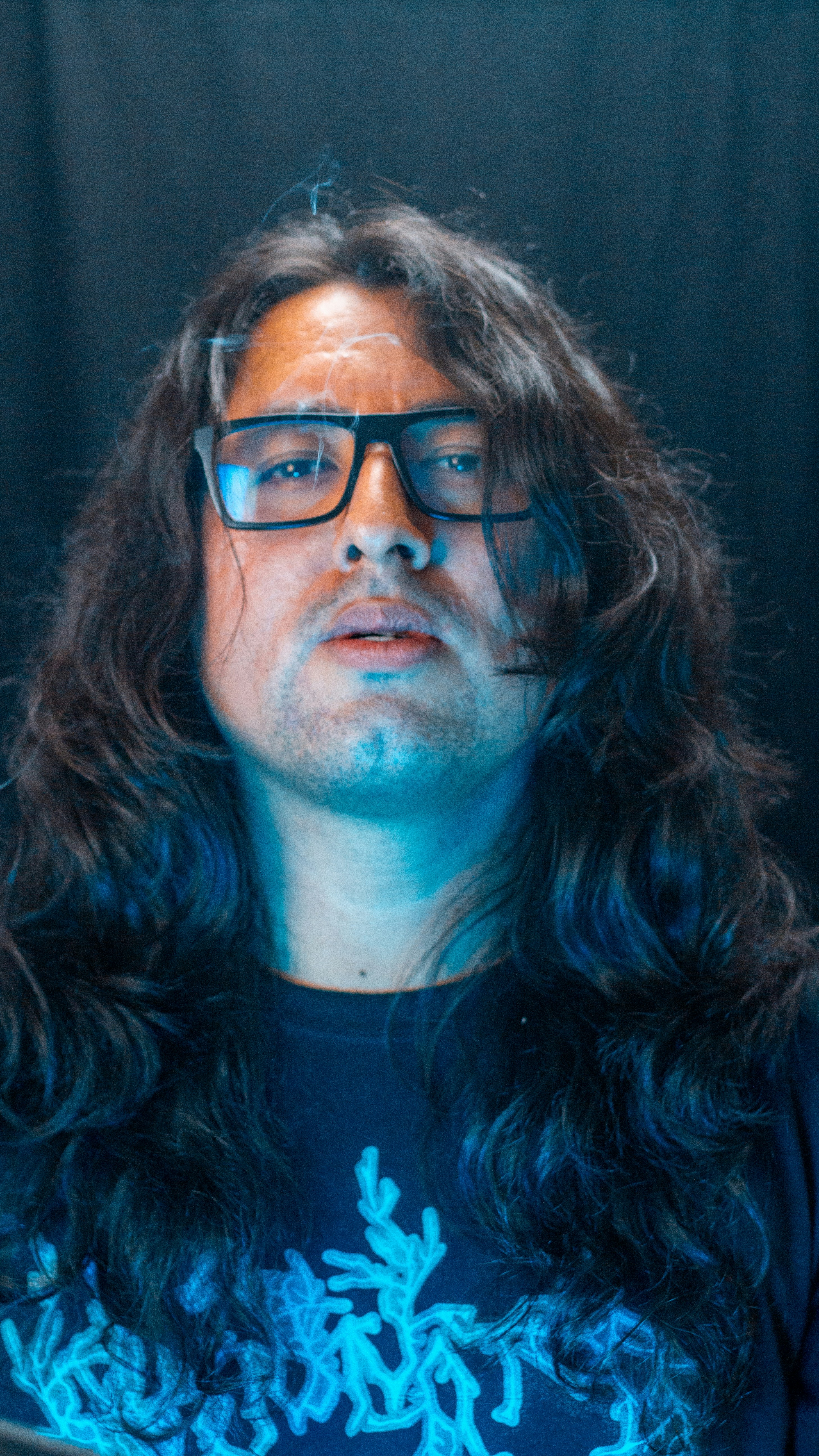 Man with long hair and glasses wearing a dark shirt.