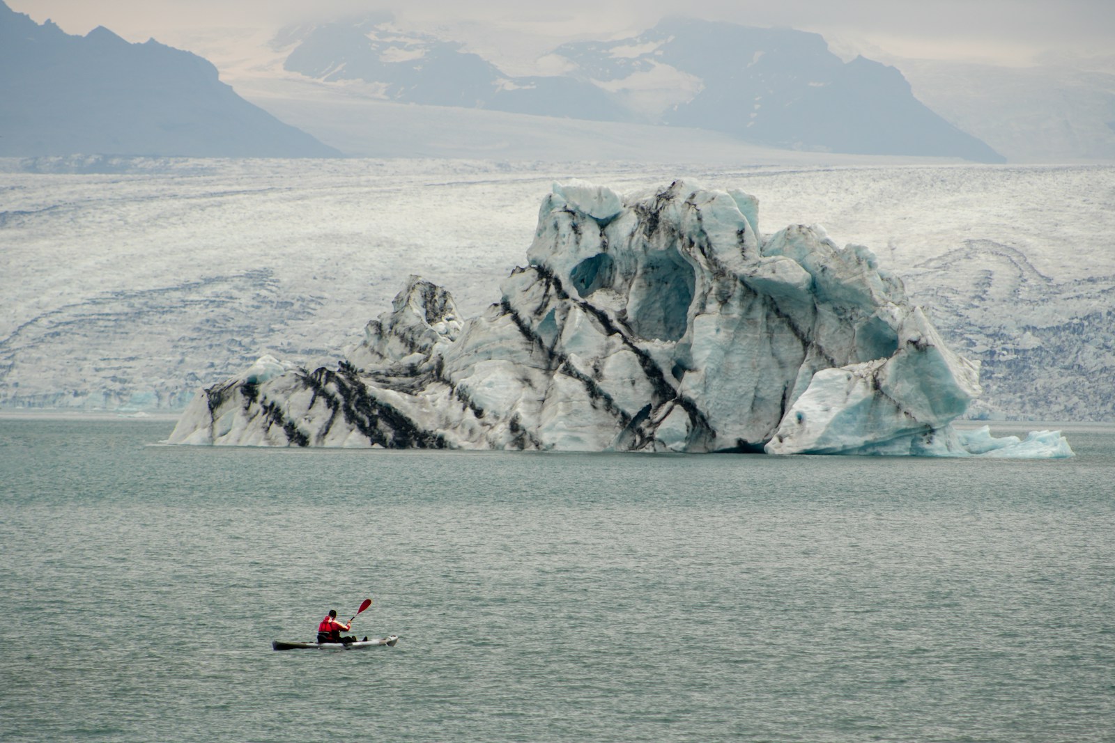 A lone kayaker in a yellow boat paddling past a black-streaked iceberg, against a wide glacier front