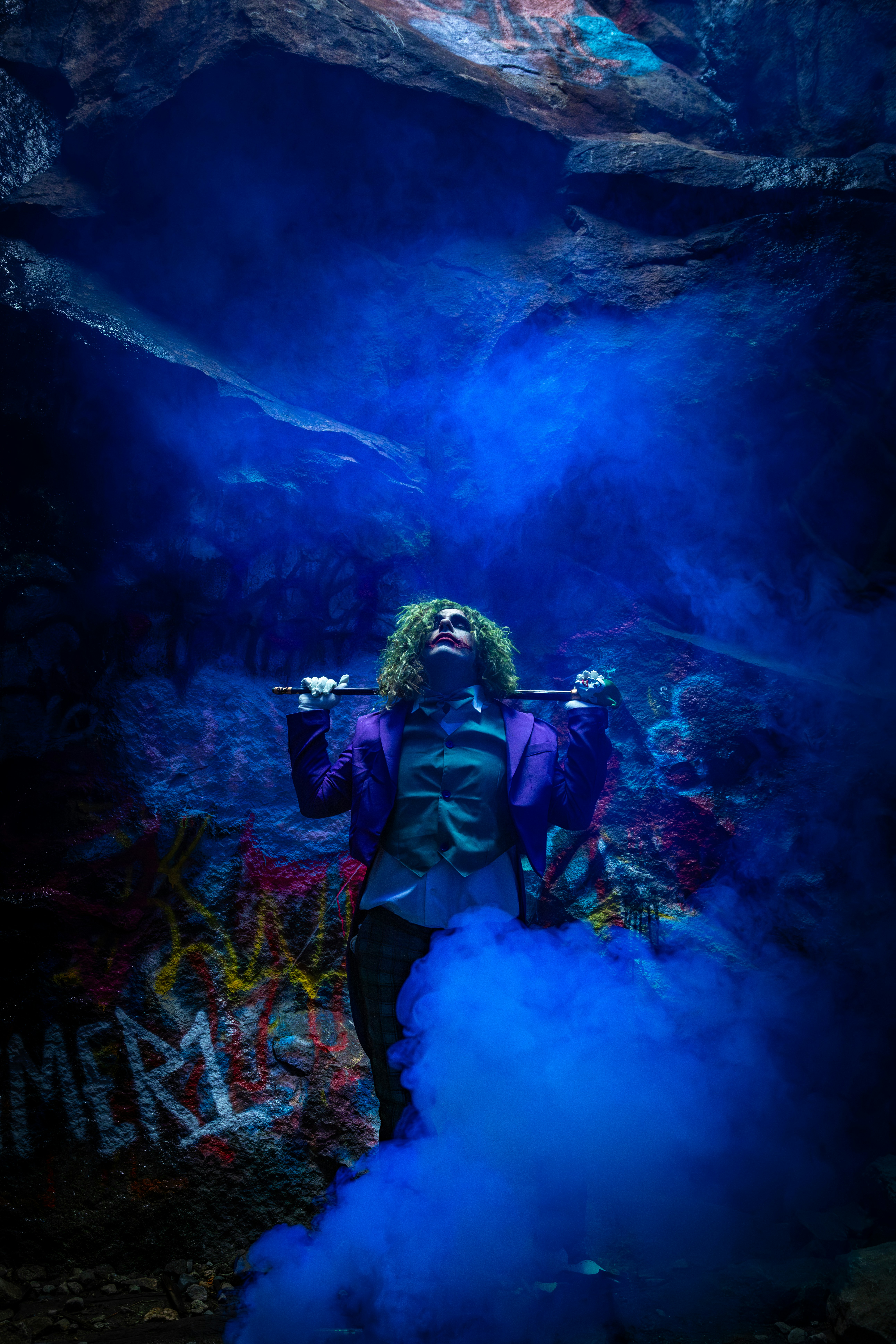 Man in joker costume with blue smoke in cave.