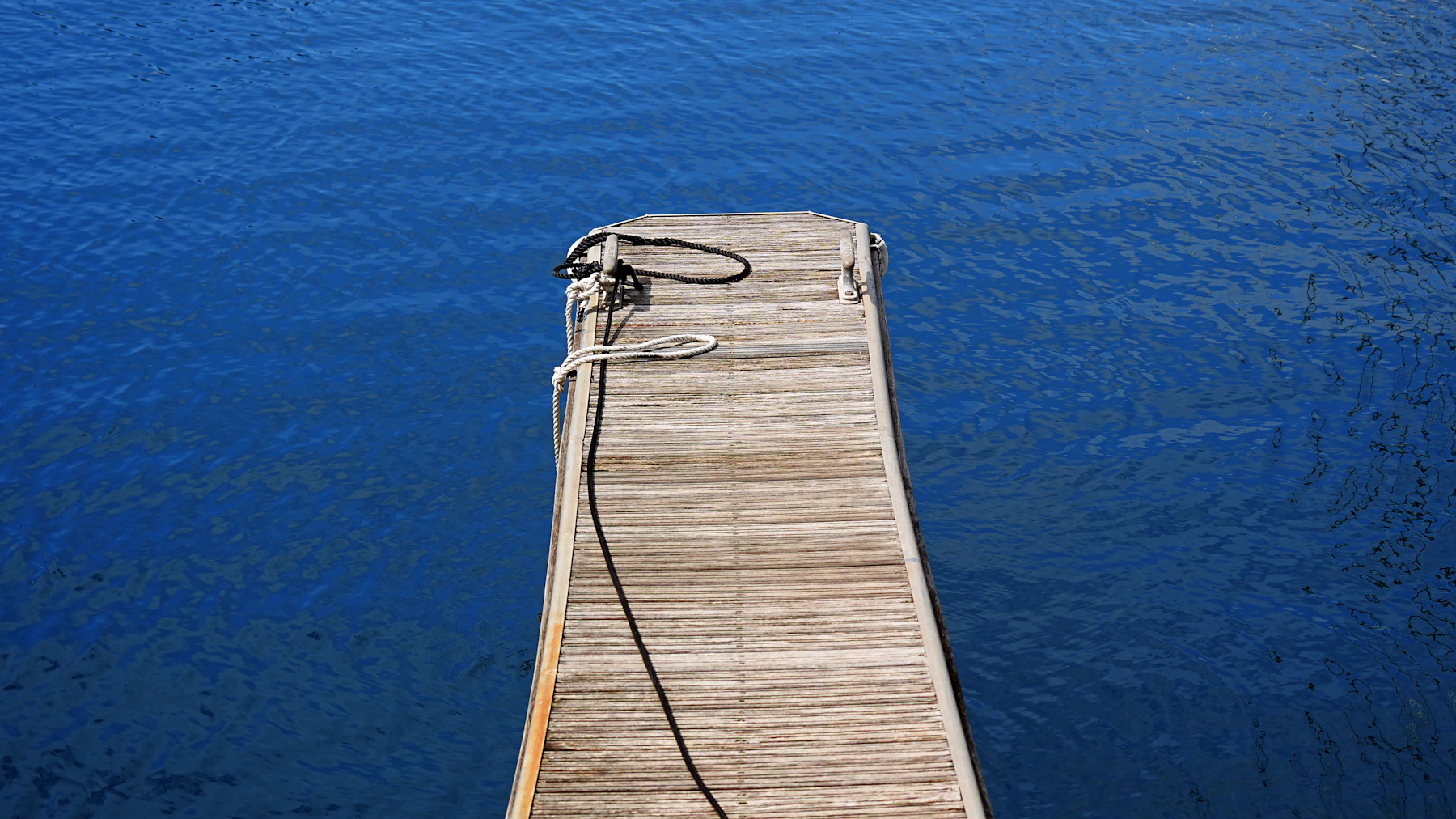 A weathered teak dock running out into deep blue water, a mooring line coiled at its end
