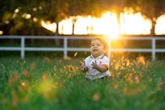 A happy baby sits in a grassy field at sunset.