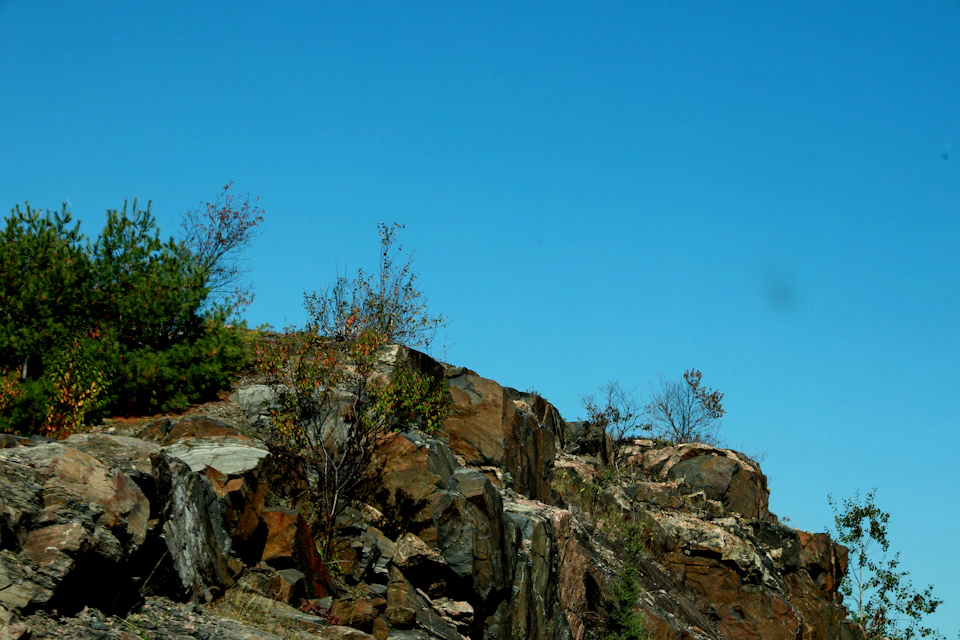 Rocky cliff face with greenery, situated on a hillside, viewed from below against blue sky