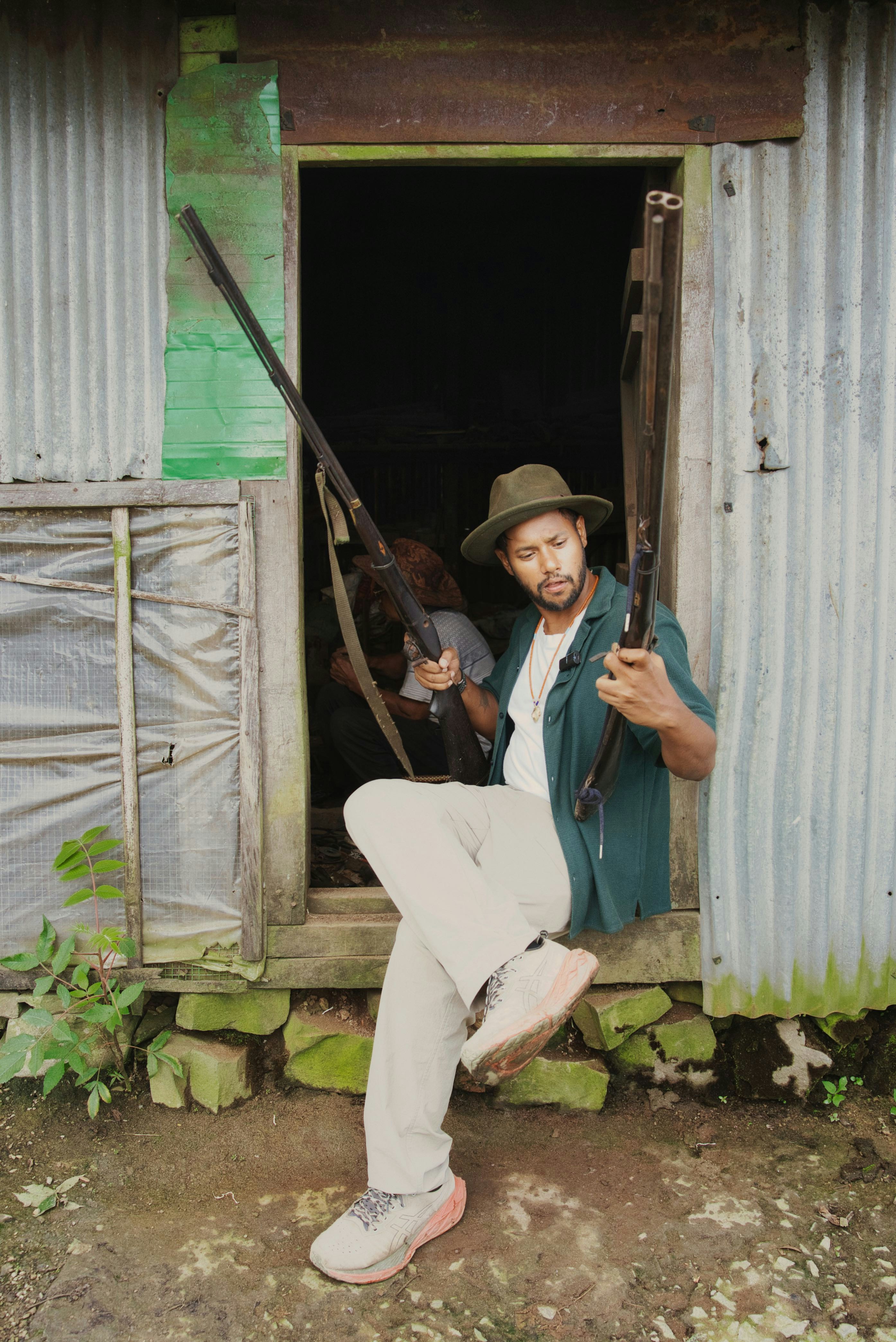 Man in hat holding two rifles by rustic doorway.