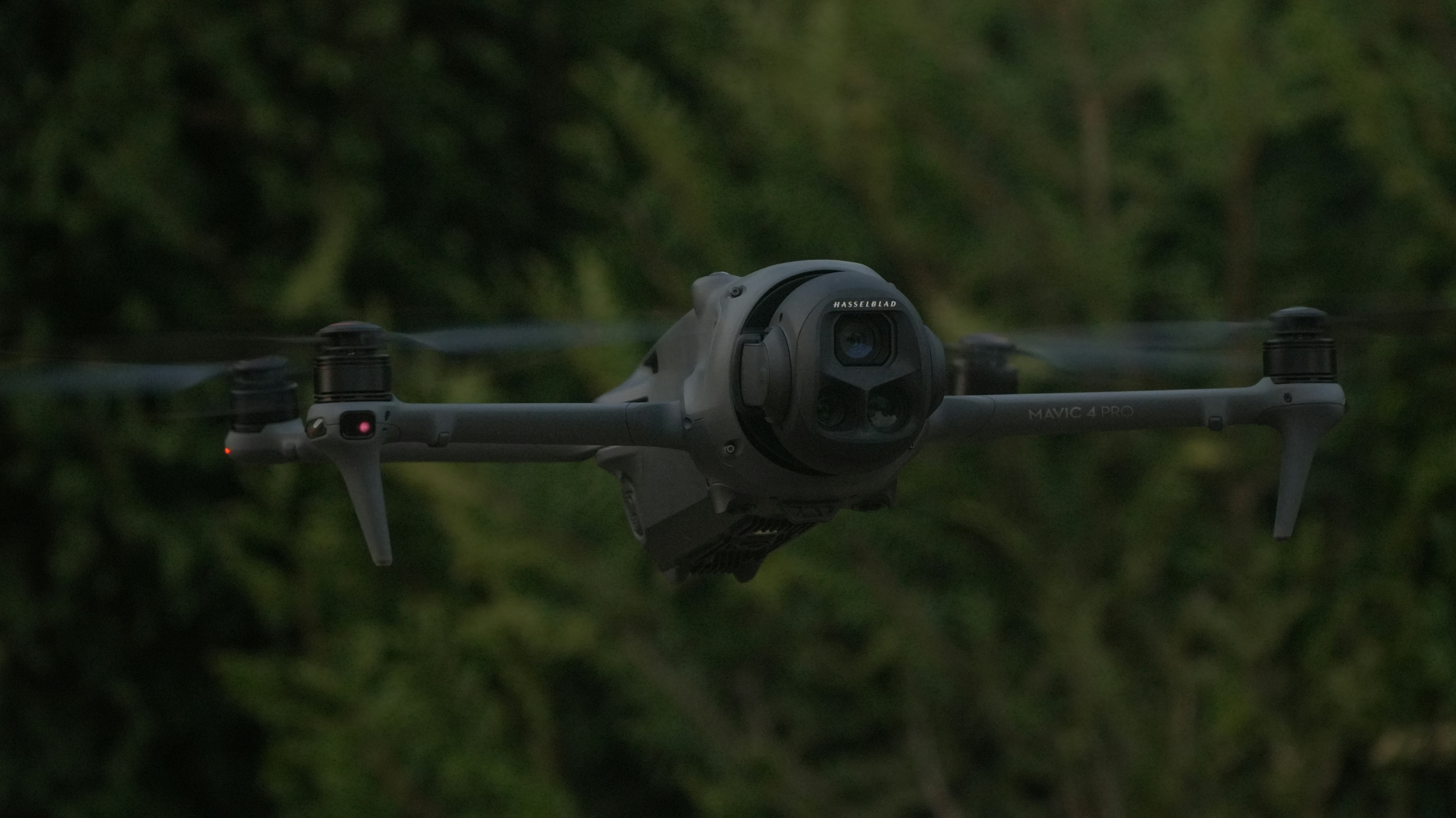 A dark drone flies in front of green trees.