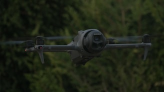 A dark drone flies in front of green trees.