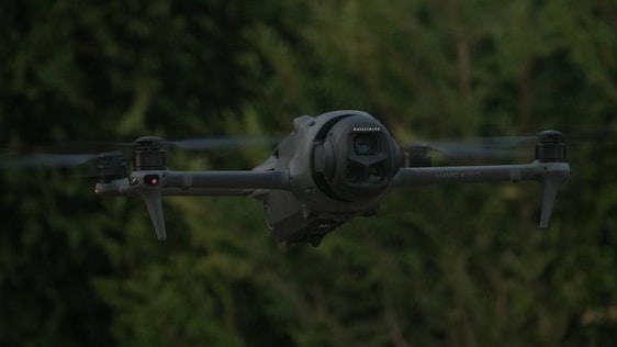 A dark drone flies in front of green trees.