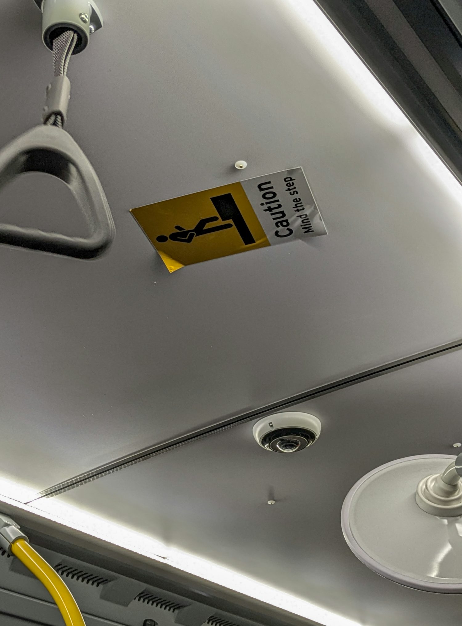 A sticker with an illustration of a person stepping with the text 'Caution. Mind the step' adhered to the roof of a bus. Also seen are handles to stablise oneself and a camera.