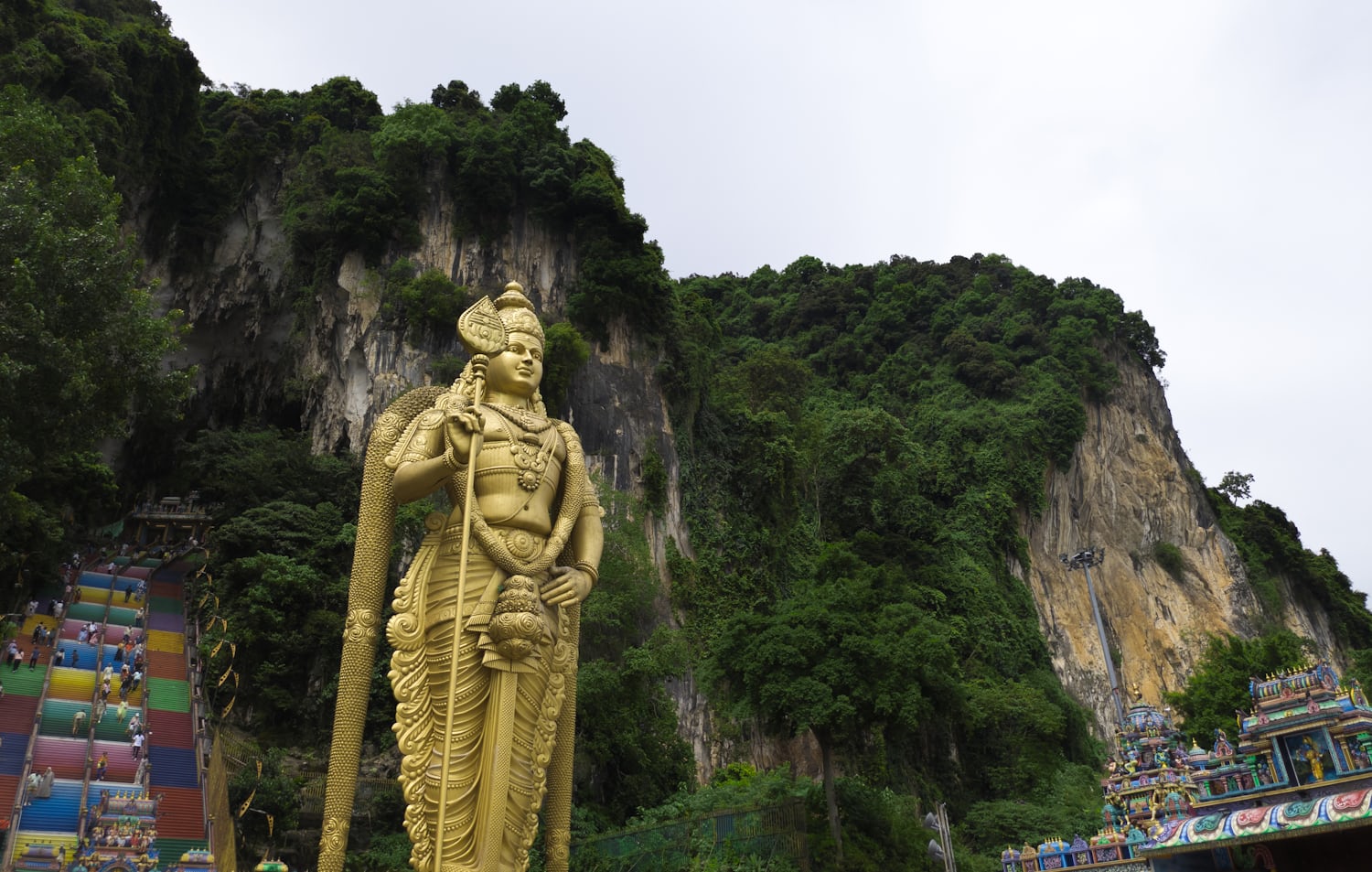 A large golden statue of a Hindu god standing against a cliff lush with green foliage. To the left of frame are rainbow steps leading to the entry of the cave, and to the right is the top of a temple.