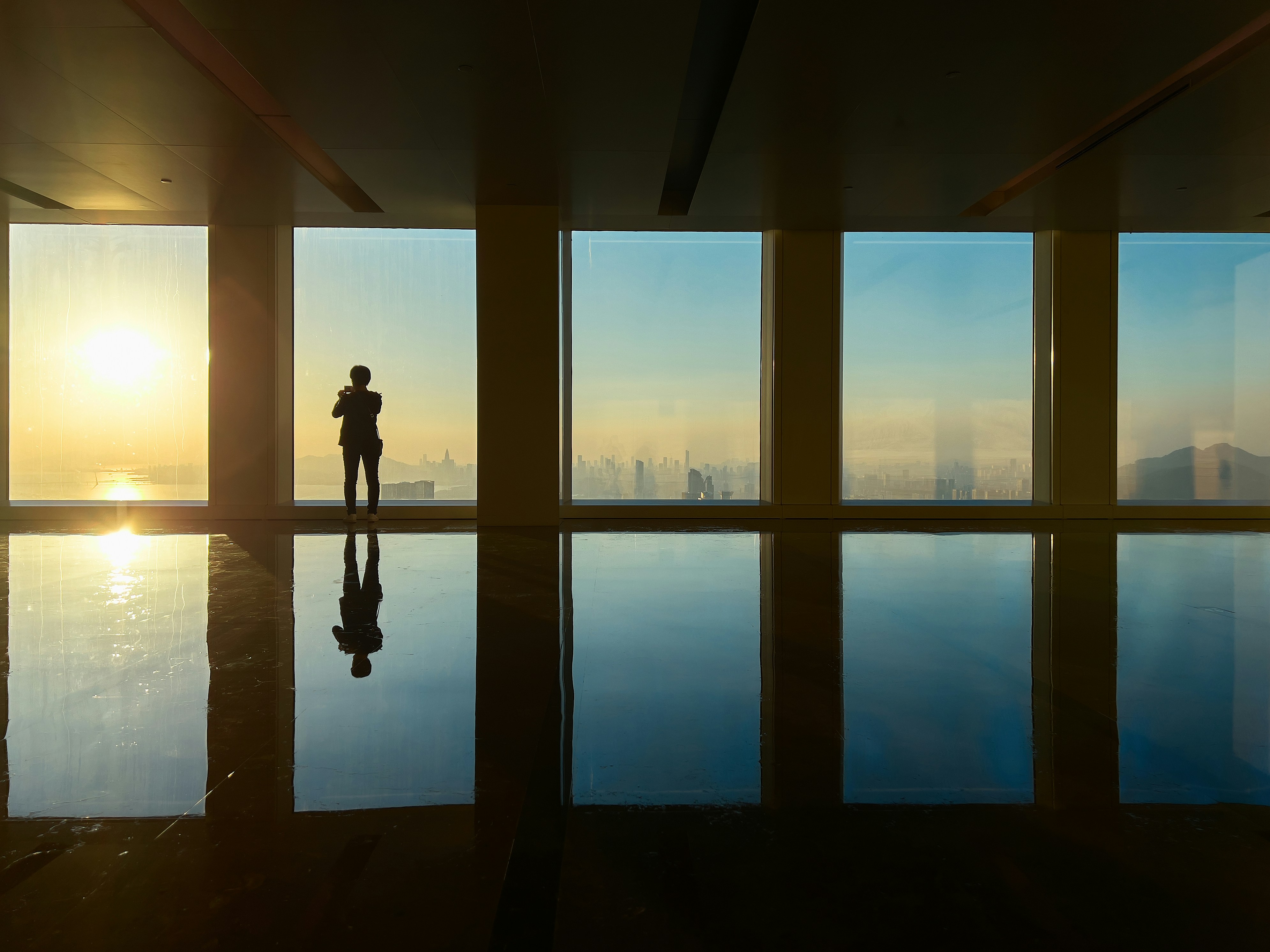 Man looking out large window at sunrise over city