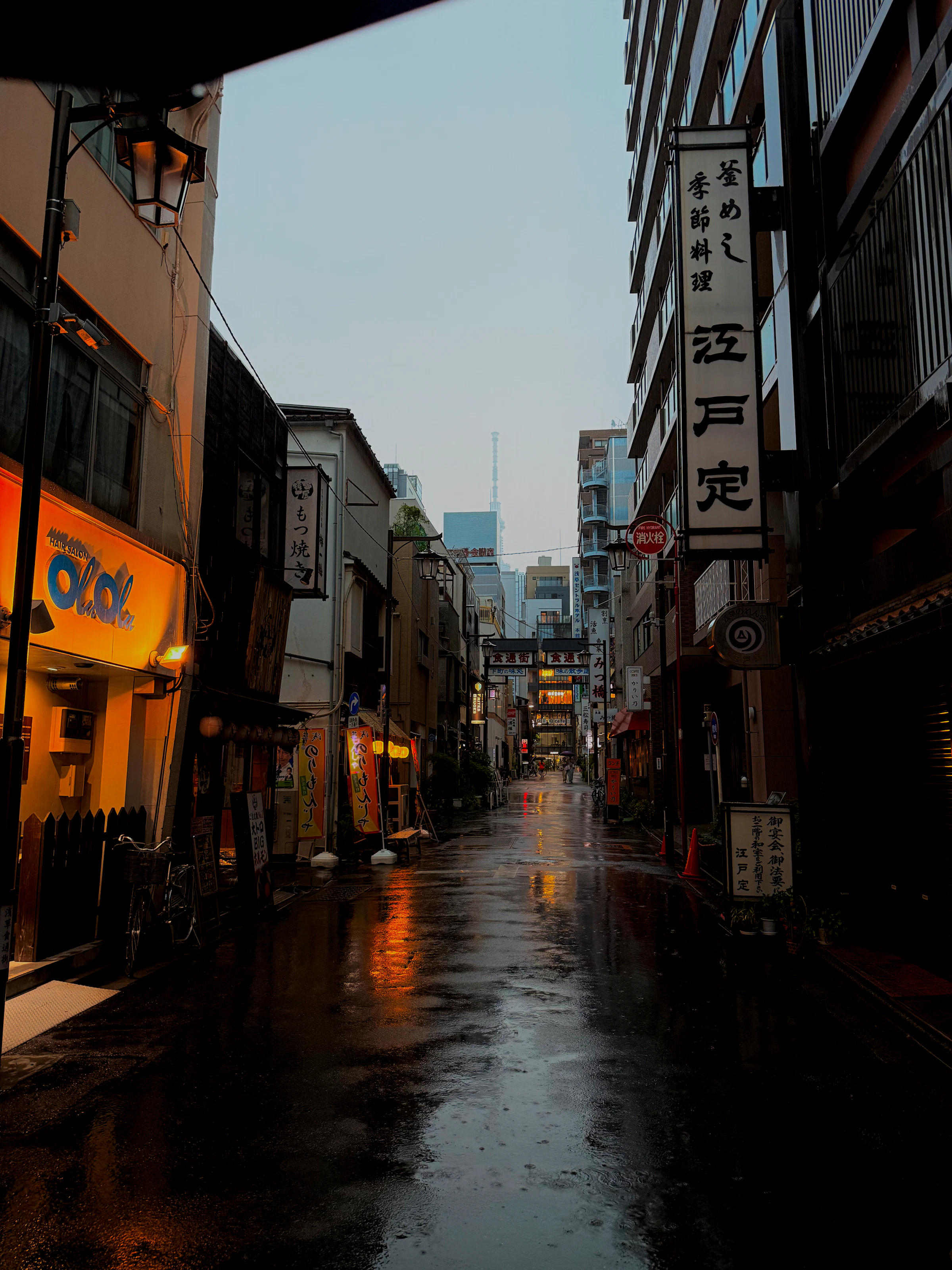 A narrow street at dusk, wet stone glowing under shop signs